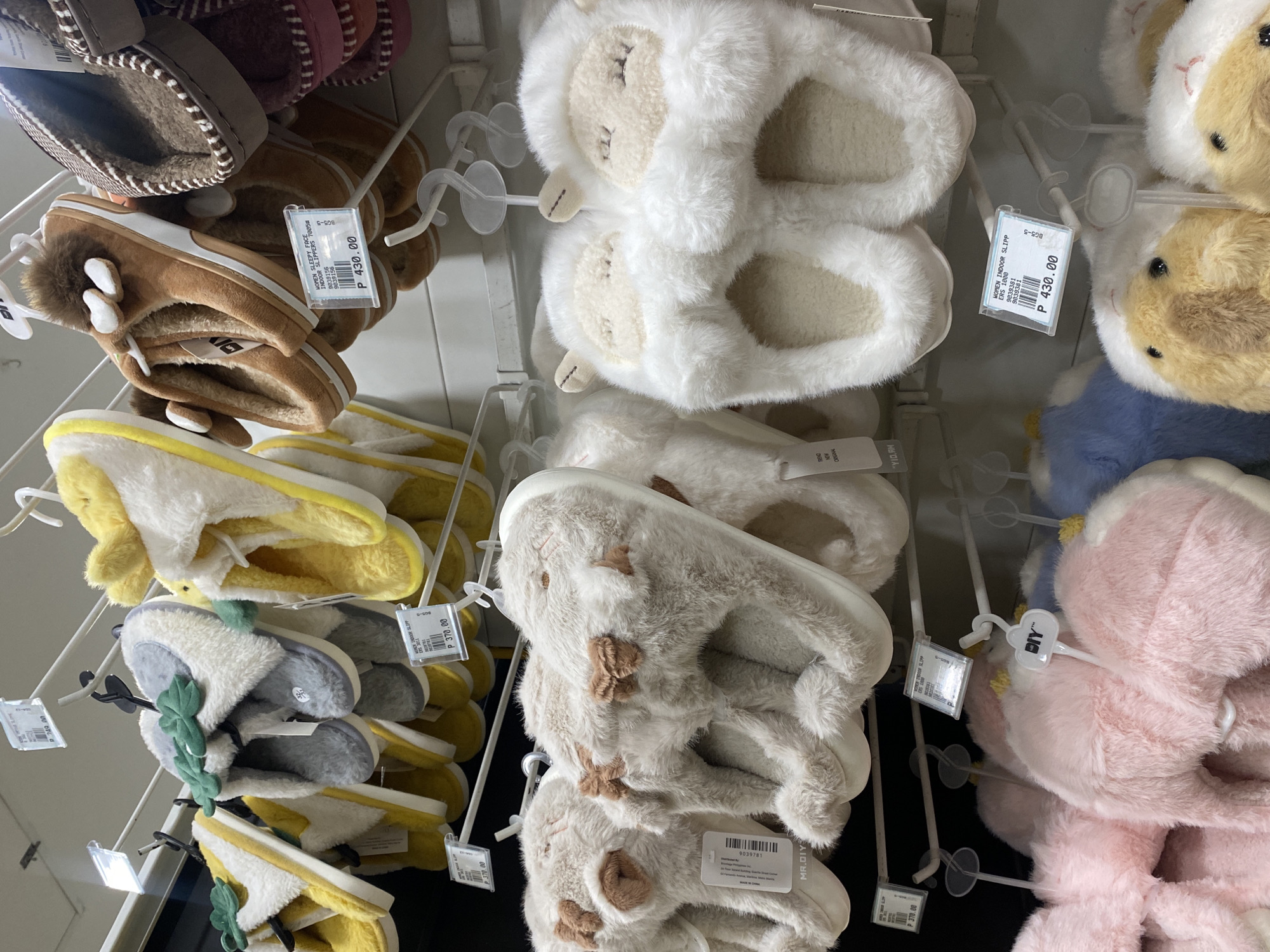 The image captures a vibrant, well-stocked display of fluffy indoor slippers in a retail store, likely a Mr. DIY branch in Davao City, Philippines. Numerous pairs of soft, comfortable slippers are neatly arranged on white wire shelving. The slippers come in a variety of designs and colors, with many featuring cute animal or cartoonish motifs.
Notable designs include white slippers with embroidered sleepy faces, light beige slippers with brown patches, tan/brown slippers with small white ears and fluffy tails, yellow and white slippers, some adorned with green leaf-like embellishments, and pink and blue fluffy pairs. Most slippers are plush and appear warm, suggesting they are designed for indoor comfort.
Price tags are visible on many items, indicating prices of "P 430.00" and "P 370.00," which denotes Philippine Pesos. Some tags explicitly state "WOMEN SLEEPY FACE INDOOR SLIPPERS" or "WOMEN INDOOR SLIPPERS." The store branding "MR.DIY" is present on several tags and a heart-shaped hanger, confirming the retail chain. Other text includes product codes like "9839156" and "9039381," as well as "MADE IN CHINA" on one label. The overall scene suggests a bright, organized shopping environment.