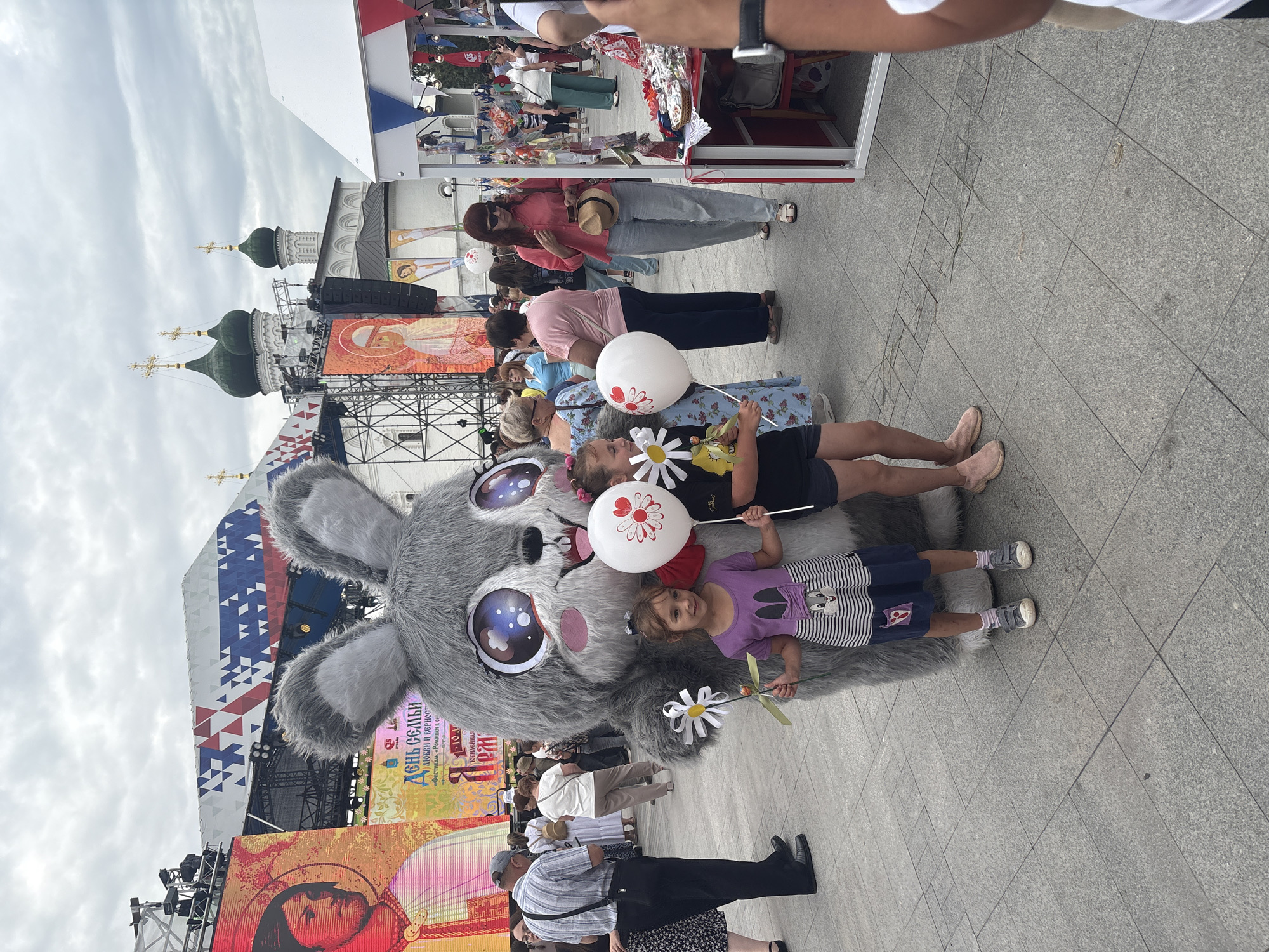 In Astrakhan, Russia, on a cloudy day, a costumed rabbit character stands on a paved plaza, posing with two young girls. The rabbit, with large, sparkly eyes and grey fur, holds a white balloon with a red flower design and a white flower prop. The girl on the left wears a purple shirt with a bunny design and a blue skirt, while the girl on the right wears a black shirt and shorts. Both girls hold similar white flower props. In the background, a large stage structure is visible, adorned with colorful banners and screens displaying portraits. Behind the stage, traditional Russian architecture with green domes and crosses can be seen. Other people mill about the plaza, with some browsing stalls set up along the right side of the image. The overall scene depicts a lively outdoor event or festival.