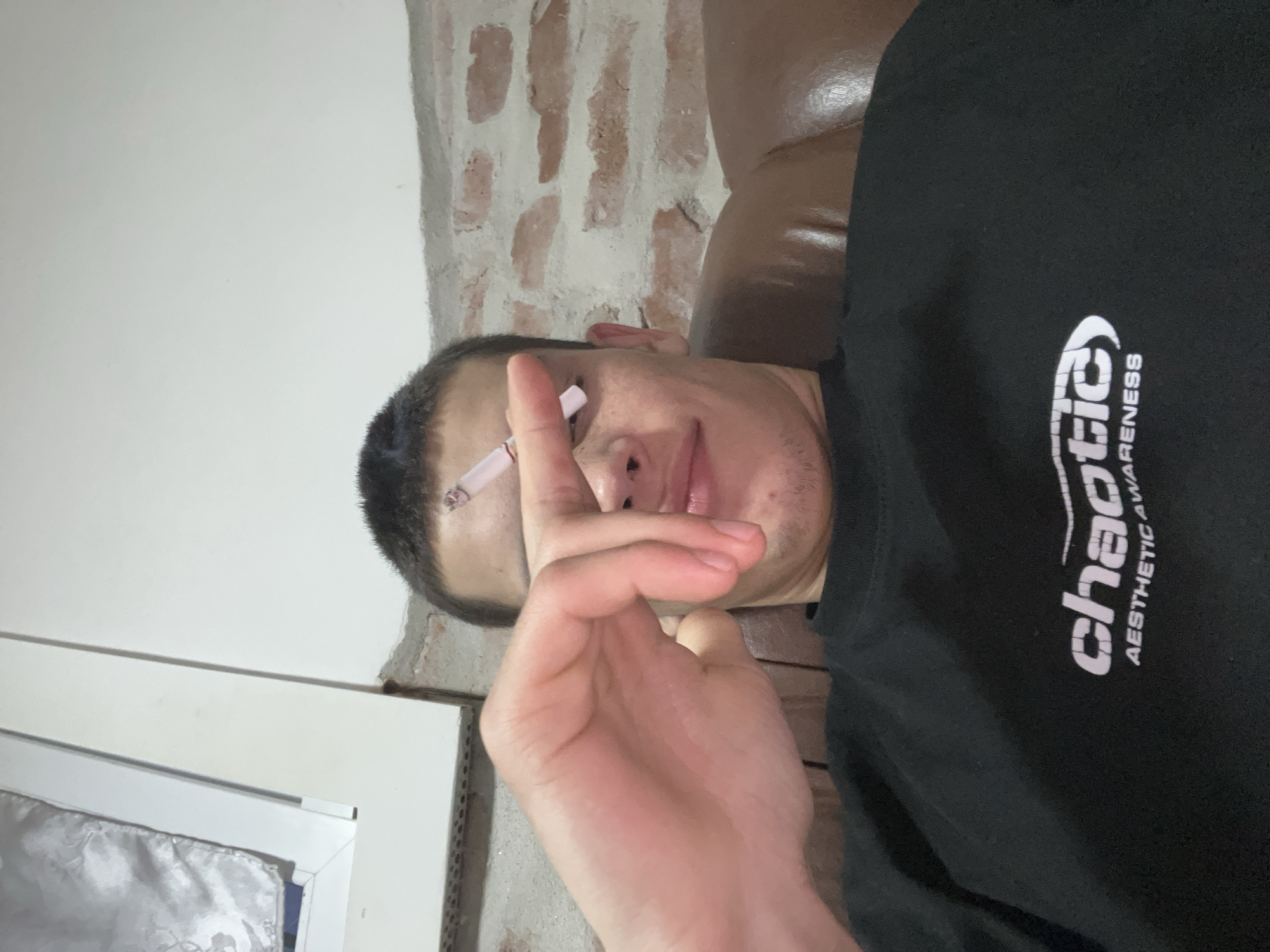 The image shows a man reclining on a brown leather sofa. He is wearing a black sweatshirt with the word "chaotic" and "aesthetic awareness" printed on it. He is holding a lit cigarette between his fingers near his face. The background includes a white wall with a window frame and what appears to be a brick wall section. The exact location in Plovdiv, Bulgaria cannot be determined from the image.