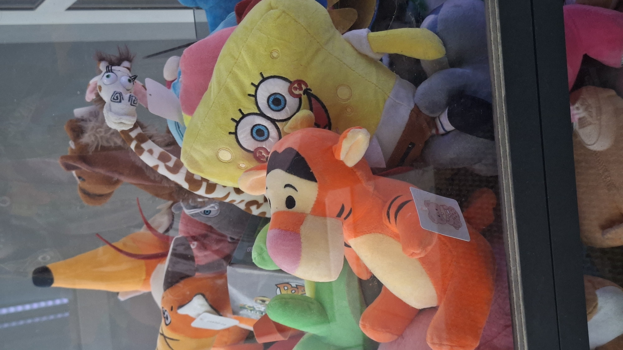 In the city of Smalyavichy, Belarus, an arcade claw machine is filled with plush toys. In the foreground, a Tigger plush toy is positioned on its side, facing left. To its right and slightly behind it, a Spongebob Squarepants plush toy is visible, with its characteristic square body and large eyes. Several other plush toys, including what appears to be a giraffe with a long neck and a brown bear, are partially obscured behind Tigger and Spongebob. The toys are packed tightly within the glass enclosure of the claw machine, with the claw mechanism visible above them. The background is indistinct and out of focus, suggesting a typical arcade environment.