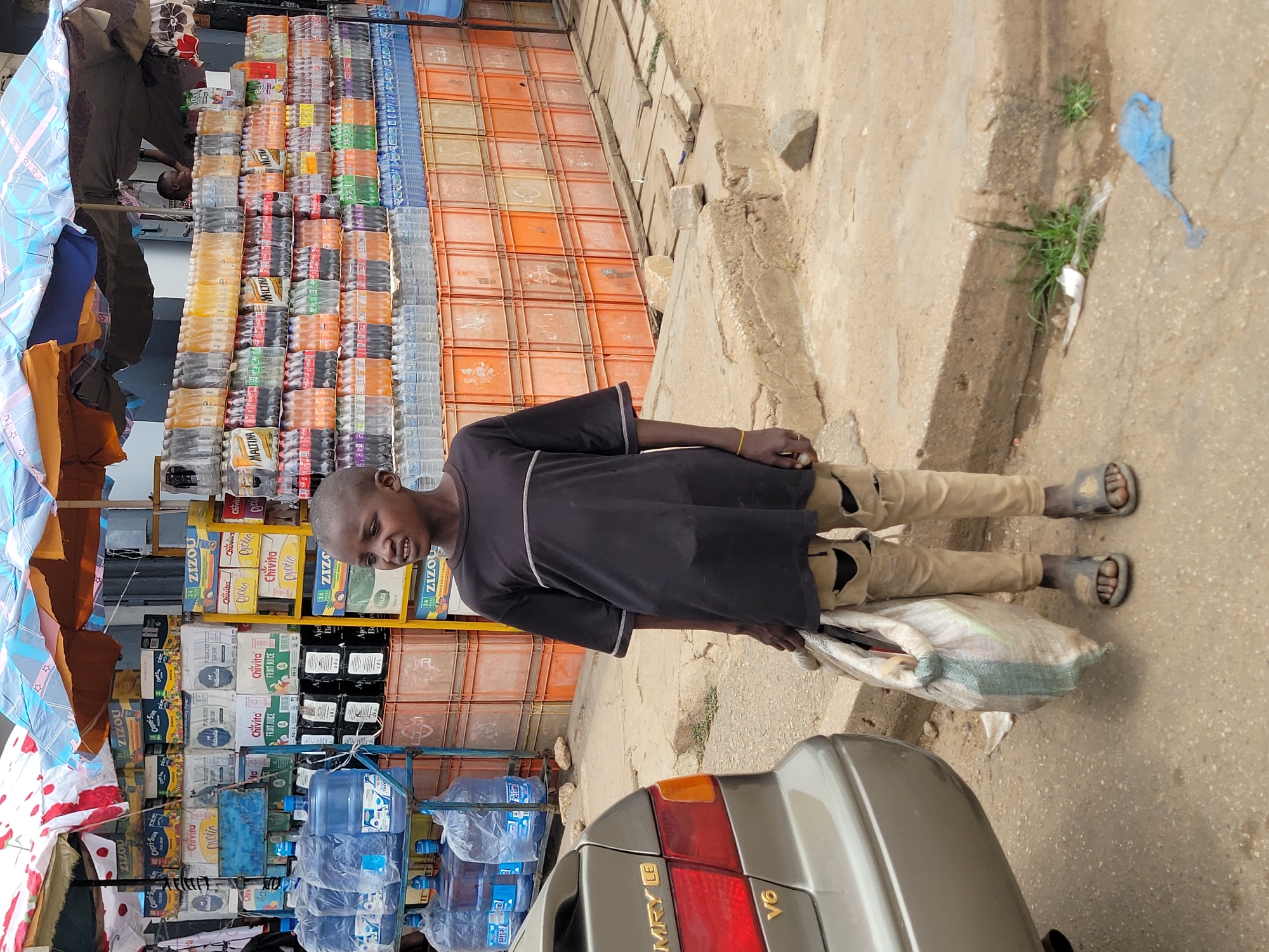 The image depicts a young boy standing on a sidewalk in front of a shop selling various beverages, including bottled water, soft drinks, and juice boxes, in Jos, Nigeria. The boy is wearing a dark short-sleeved shirt with a white stripe, tan pants, and sandals. He is holding a partially filled white sack. A portion of a silver car is visible in the foreground. The shop displays rows of stacked drinks and beverages, with visible brands such as Coca-Cola, Fanta, ZIZOU, Chivita, and Maltina. The street environment includes a concrete curb, some vegetation, and scattered debris. The image is taken in daylight, with natural lighting.