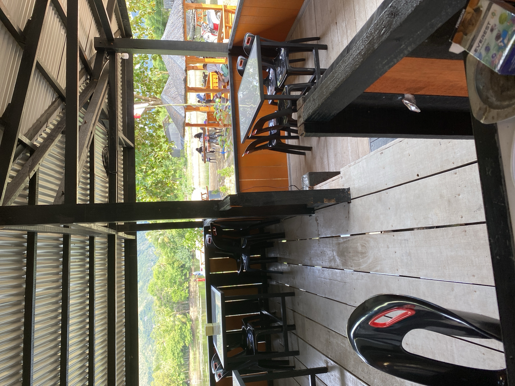 The image depicts an outdoor dining area with tables and chairs situated on a wooden deck. The area is covered by a corrugated metal roof supported by dark-colored posts. Beyond the deck, there is a body of water surrounded by lush green trees and vegetation, with hills visible in the background under a clear sky. Several thatched-roof gazebos are present near the water's edge. People are seen seated at tables in the gazebos and near the water. The foreground shows a black chair and a portion of a table. In the lower right corner, a pack of cigarettes and an ashtray are visible. The overall scene suggests a relaxed, possibly tropical, waterfront dining establishment.