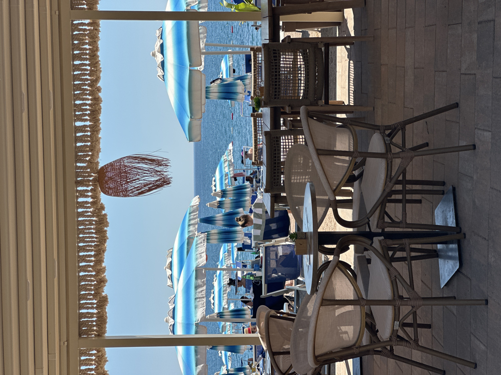 The image shows a seaside scene, likely a beach resort or café, in Boreti, Montenegro. Rows of blue and white striped beach umbrellas line the shore, seen through the frame of a covered patio. The patio features a woven ceiling decoration and furnishings including several woven chairs and small tables. The focus is on the patio furniture in the foreground. Beyond the beach umbrellas is the sea and a glimpse of the horizon. The sky is clear and blue. People are visible under the umbrellas on the beach.