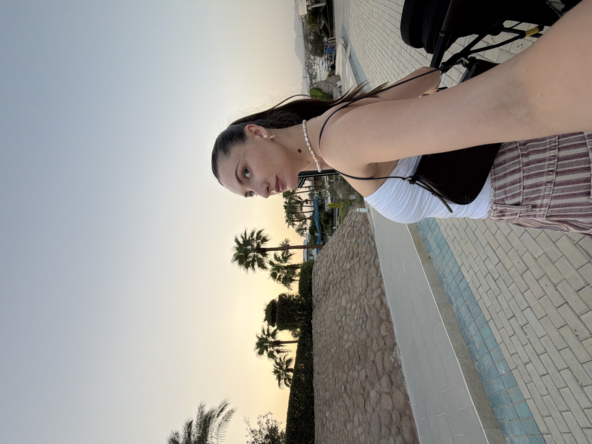 In Sharm El Sheikh, Egypt, a woman is captured in a selfie-style photograph, walking on a paved pathway. She is wearing a white tank top, a striped skirt, and a dark handbag slung across her shoulder. A pearl necklace adorns her neck, and she has on small earrings. The background features palm trees, a stone wall, and distant mountains, suggesting a resort or outdoor area with a clear sky transitioning to dusk.