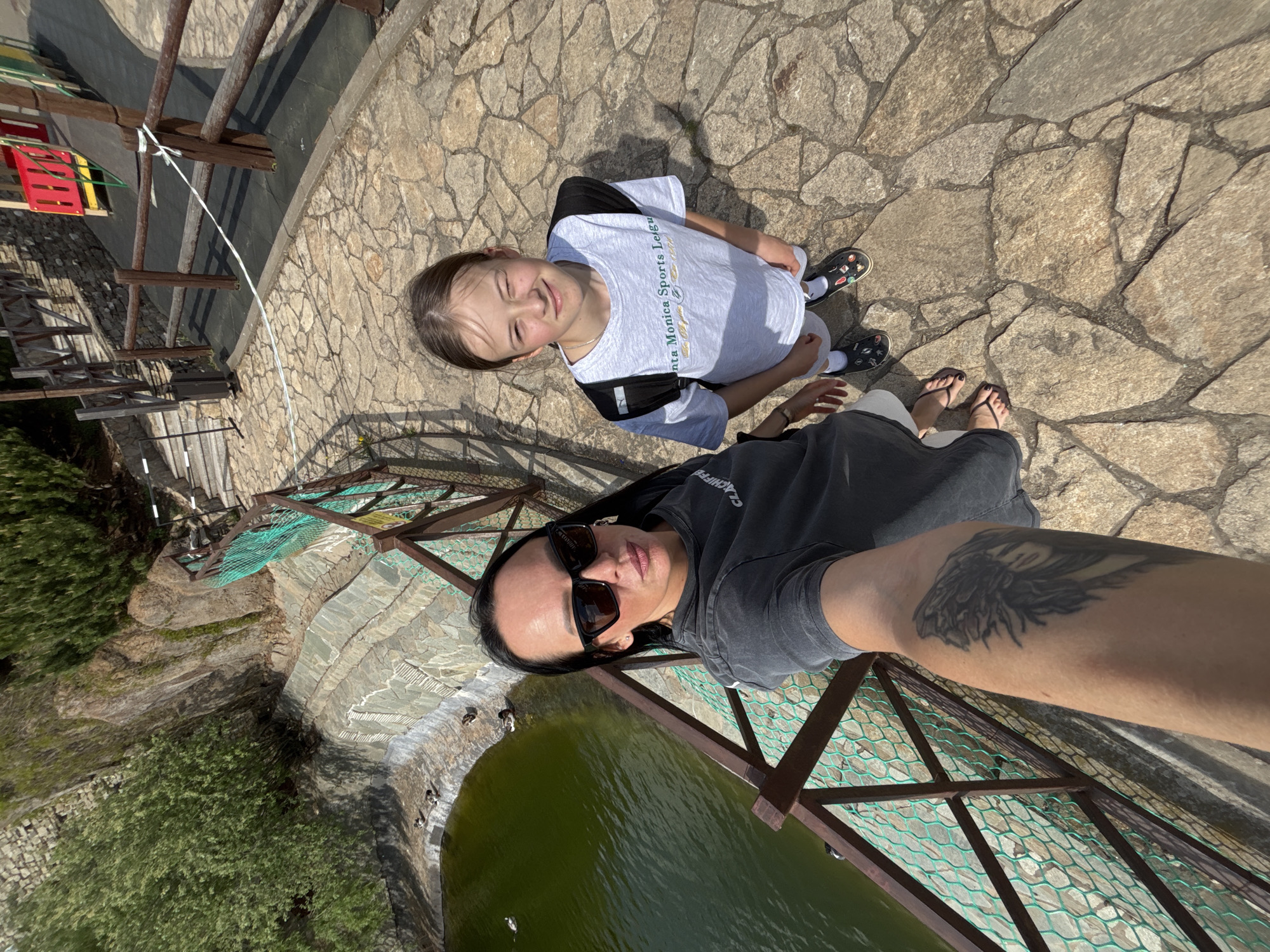 A woman with sunglasses and a tattoo on her left arm is taking a selfie with a young girl on a stone surface. The woman is wearing a grey shirt and flip-flops. The girl is wearing a white t-shirt with "Santa Monica Sports League" printed on it and black and white Crocs.
They appear to be standing on a bridge or platform with a metal railing, overlooking a body of water, potentially a pond, surrounded by trees and greenery. The setting includes stone pathways, wooden structures resembling fences or playground equipment, and possibly a retaining wall made of stacked stones.
The photograph seems to be taken in a park or recreational area. Although the exact location in Izhevsk, Russia, cannot be determined from this image alone, the presence of these features suggests it could be a park or recreational area in the city.
