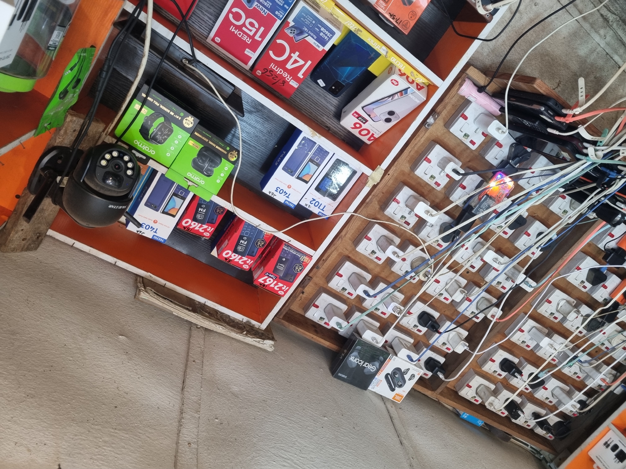 The image depicts a busy electronics stall or shop in Fegge, Nigeria, characterized by a dense arrangement of mobile phones, smart devices, and a communal charging station.
On the left, shelves are packed with new mobile phones in their packaging. Visible brands include Redmi (models 15C and 14C, showcasing specifications like 8GB RAM 128GB ROM and 16GB RAM 256GB ROM), Itel (A06, it2160, it2167), and Tecno (T403, T102). Price tags such as "15k", "26k", and "17.5k" (likely denoting thousands of Nigerian Naira) are visible on several phone boxes. Other merchandise includes a "SOLID STAR" brand security camera, "oraimo" smartwatches and wireless earbuds (with text like "2.01" ND SCREEN, FULL CONTROL" and "IMMERSIVE SOUND, EVERYWHERE"), "JBL BUDS," and "SAMSUNG Gear Iconx V5.0."
The right side of the frame is dominated by a custom-built wooden board housing numerous electrical sockets. A multitude of power adapters are plugged in, creating a dense network of multi-colored charging cables (white, blue, black, pink, orange) that extend from the board. These cables are actively charging various mobile phones, some visible and others implied to be off-screen. A small pink flashlight and a green plastic pouch are also present near the charging area. The scene conveys a bustling commercial activity focused on selling and charging electronic devices.