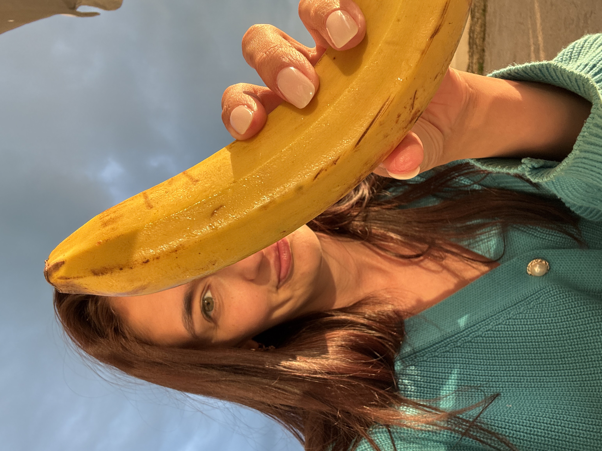 A woman with shoulder-length brown hair and green eyes holds a ripe banana in front of her face. Her nails are painted a light pink. She is wearing a turquoise sweater with a pearl button. The sky, visible in the background, is overcast with gray clouds. The location of the photo is unknown and cannot be determined from the image.