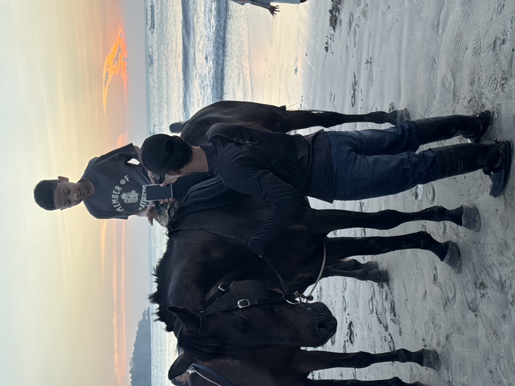 An over-the-shoulder shot captures a boy on horseback at sunset on Kuta Selatan beach, Bali, Indonesia. The boy, wearing a dark t-shirt with a graphic print, sits atop a dark-colored horse, holding a mobile phone. The horse is equipped with a saddle and bridle. A person in a black long-sleeved shirt, dark jeans, and black boots stands beside the horse, appearing to assist the boy. The sandy beach is wet from the receding tide, with footprints visible. In the background, the ocean waves gently roll ashore under a sky painted with hues of orange and pink from the setting sun. A few boats are visible on the horizon. The overall scene suggests a leisurely moment at dusk on the beach.
