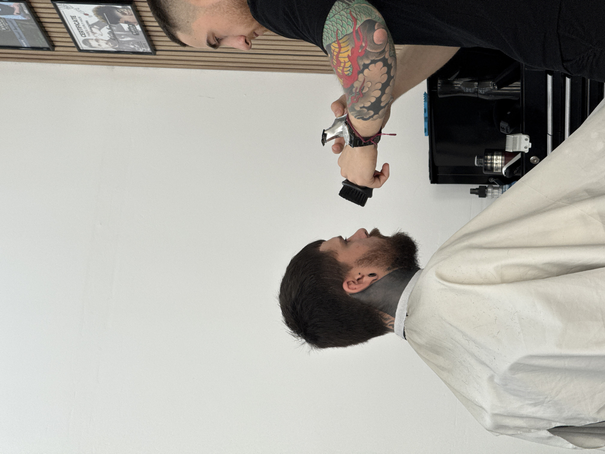 This image captures a moment in a barbershop in Sofia, Bulgaria, depicting a barber attending to a client. The image is oriented vertically, with the subjects appearing rotated to the side.
The barber, positioned at the top left when correctly oriented, is a man wearing a black short-sleeved shirt, revealing a heavily tattooed left arm featuring a vibrant, colorful dragon design. He holds a silver hair clipper in his right hand, pointed downwards towards the client, and a small black brush in his left hand. He is looking intently at the client below him.
The client, positioned at the bottom right, is a man with dark hair and a full beard. He has visible black tattoos on his neck and throat area, extending beneath his left ear. He is covered by a light-colored barber's cape, which is secured around his neck with a white strap, suggesting he is undergoing a haircut or beard trim. He is looking upwards towards the barber.
The background consists primarily of a plain white wall. In the upper left corner, several framed items, likely certificates or photographs, are mounted on a wooden panel. One frame clearly displays the word "CERTIFICATE" and other text. To the right of the barber, a black barber's station or tool cabinet is visible, holding various grooming supplies. One bottle on the station is labeled "VACUUM POWDER". The lighting suggests an indoor, daytime setting.