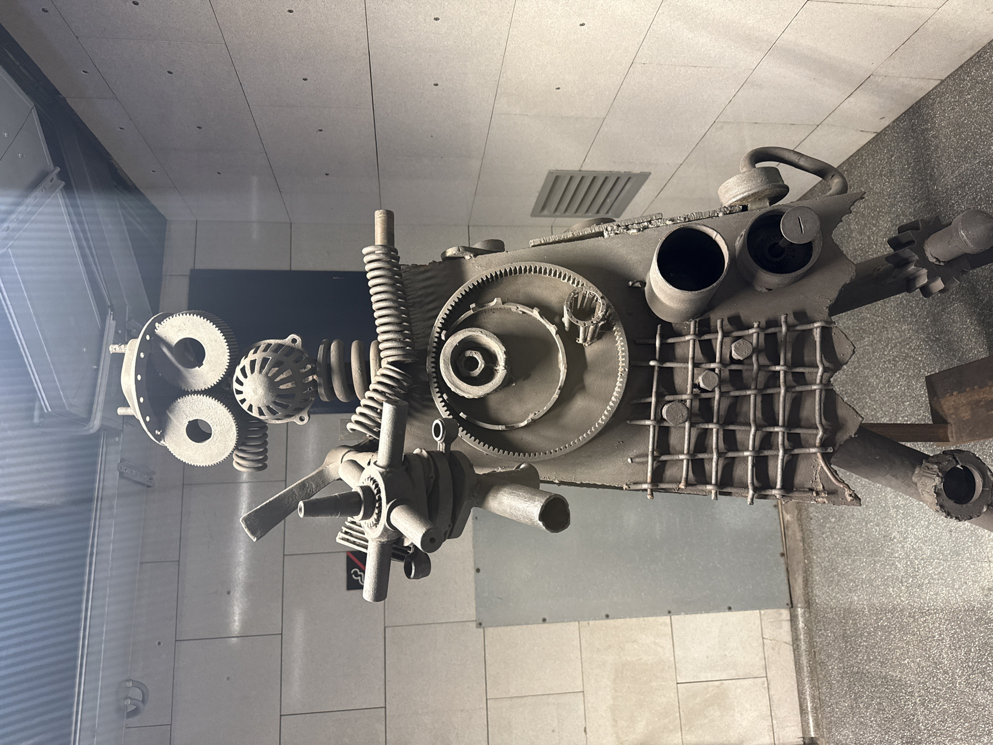 The image displays a sculpture constructed from various metal components, including gears, springs, pipes, and a grid-like structure. The sculpture appears to be an abstract representation of a mechanical form. It is located in Barcelona, Spain. The surrounding environment consists of tiled walls and flooring, suggesting an indoor setting, possibly a gallery or public space. There are no people visible in the image. The sculpture rests on a surface, and the angle of the shot suggests it may be behind a glass barrier.