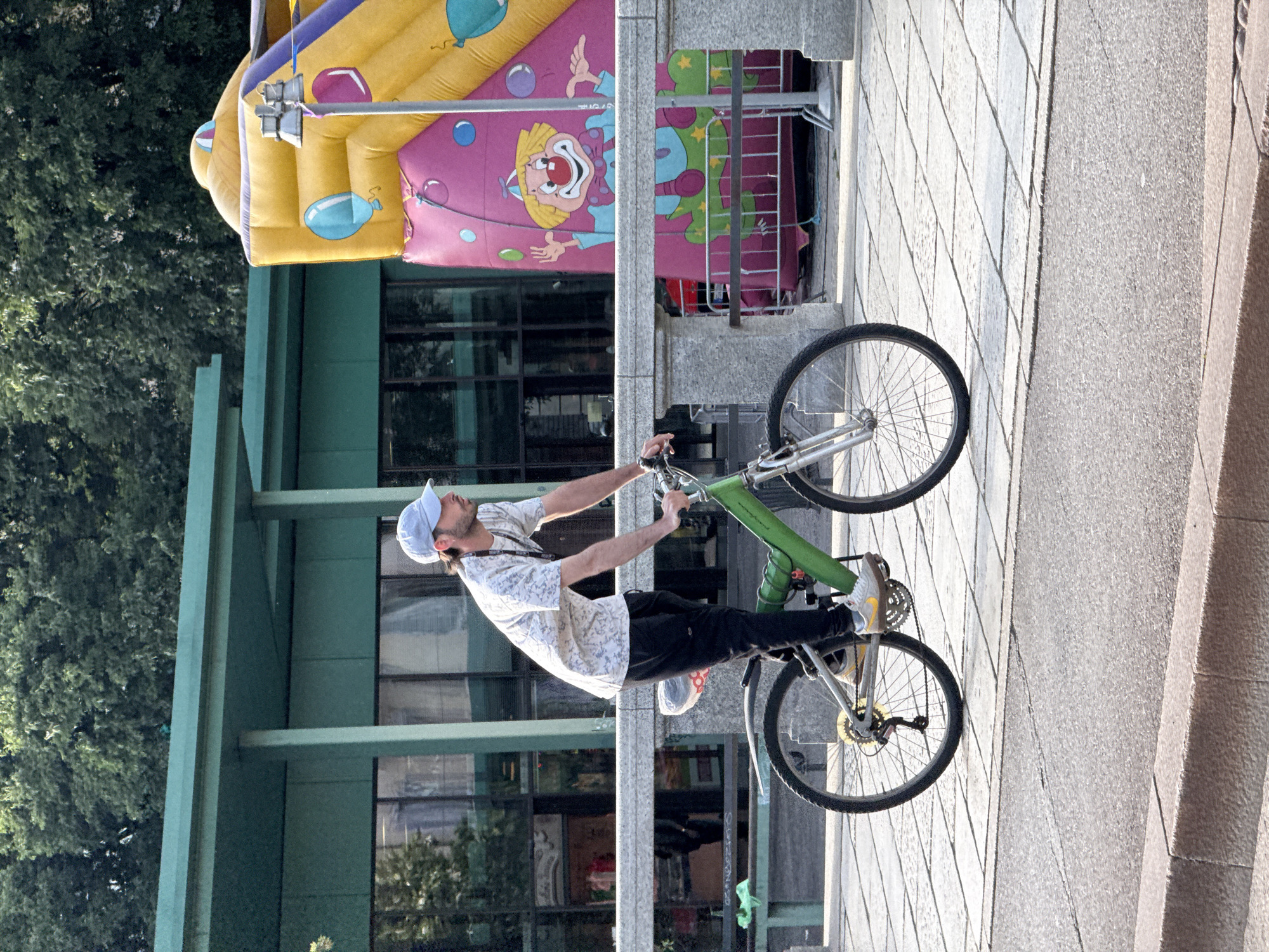 A man in a white patterned shirt, black pants, and a blue baseball cap is riding a green bicycle on a paved area. He appears to be balancing or performing a trick. Behind him is a building with green accents and reflective windows, next to a large inflatable structure featuring colorful balloons and a clown design. The background includes trees and what appears to be a cityscape. Given the style of architecture, the photograph seems to have been taken in an urban environment, but the specific location in Milan, Italy is difficult to determine without further landmarks or context.