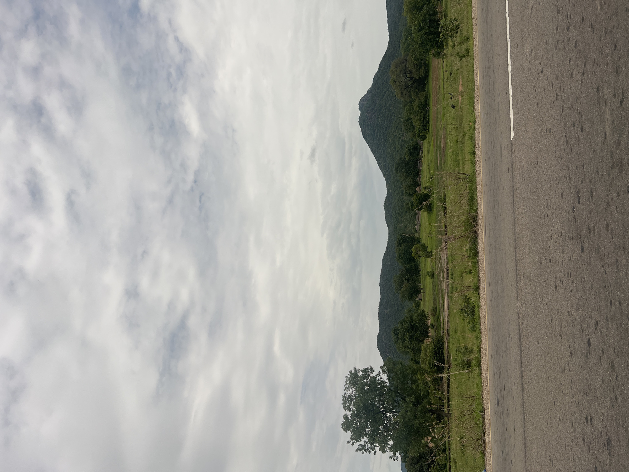 The image shows a landscape with a paved road in the foreground. Beyond the road is a grassy field with scattered trees and a lush green hill in the background. The sky is overcast with clouds. The scene appears to be rural, likely in or near Minda, Nigeria, given the description of the area. There are no people or animals visible in the image. The environment is natural and serene.