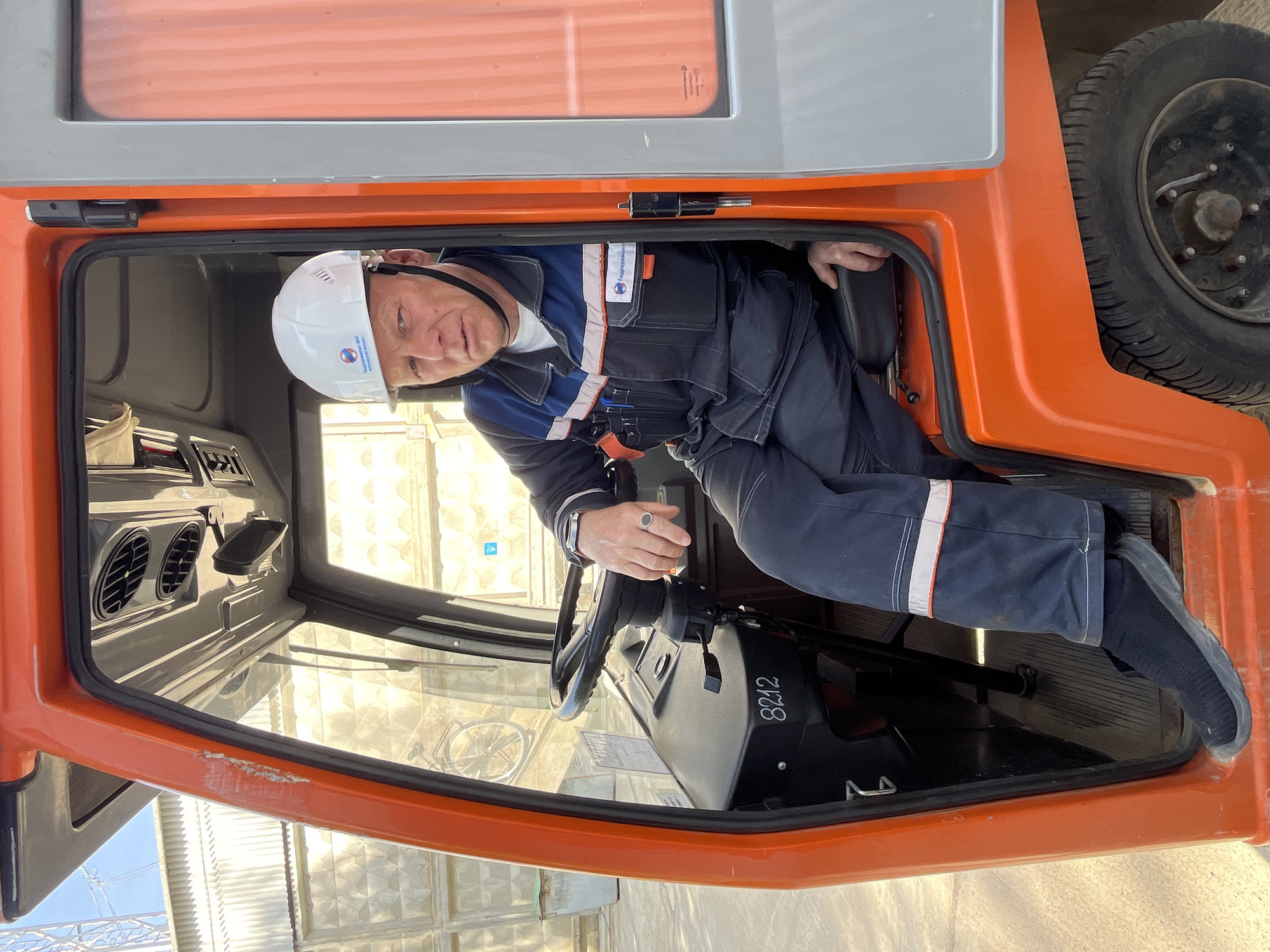 A man wearing a white hard hat and a blue and navy work uniform is sitting in the driver's seat of an orange vehicle. The uniform has a logo on the chest and white reflective stripes on the sleeves and pants. The steering wheel is black, and the dashboard displays "8212". The vehicle is parked outside, and the reflection in the window shows a building and a bicycle. The location is Chaikovsky, Russia.