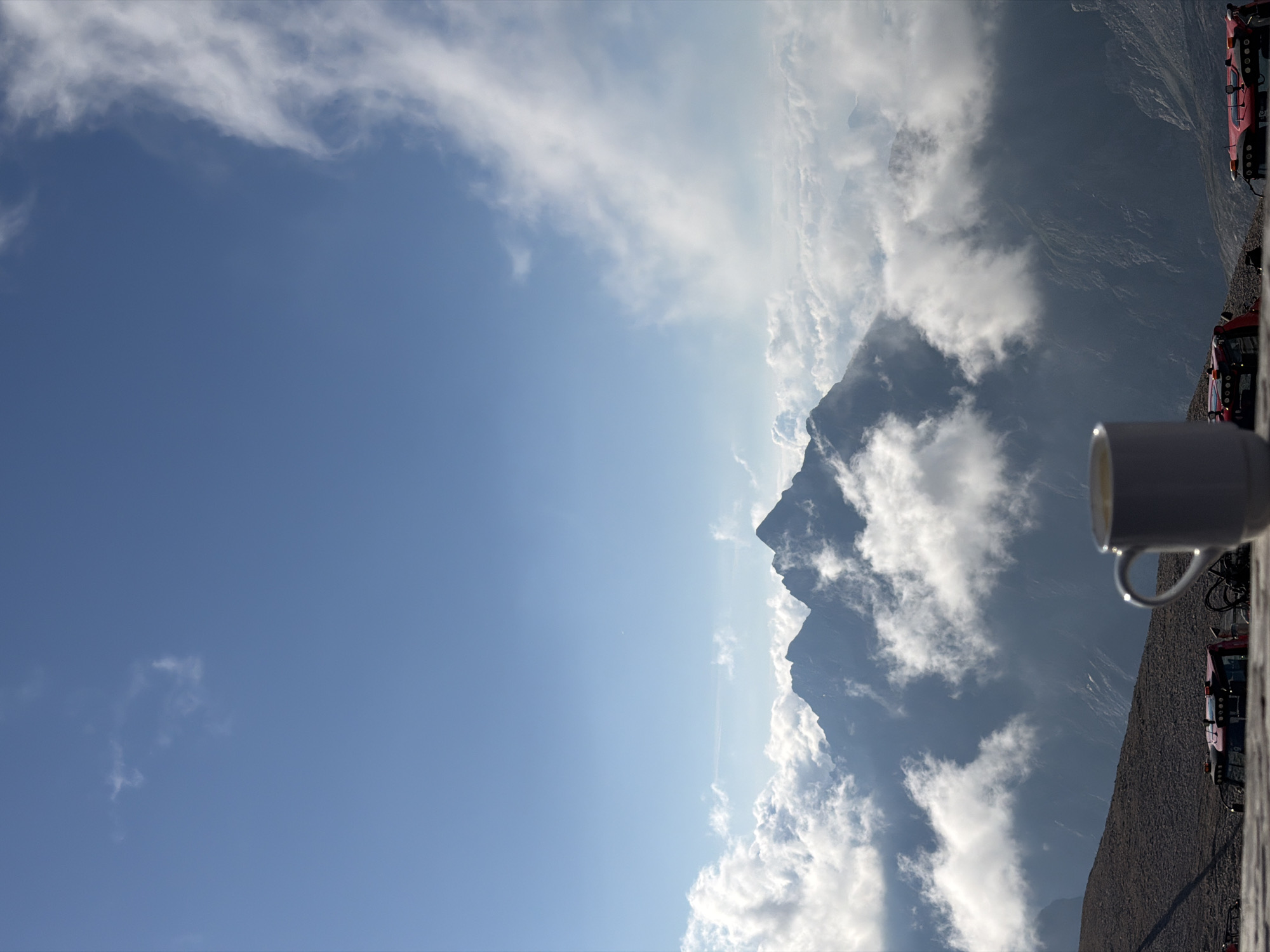 An outdoor shot features a white mug in the foreground, filled with a light-colored liquid, likely coffee or tea. Behind the mug, a vast mountain range pierces through a sea of clouds under a clear blue sky with scattered white clouds. The mountains appear rugged and imposing, with some peaks shrouded in mist. In the lower portion of the image, two red vehicles, possibly construction or utility vehicles, are visible on a rocky, uneven terrain, hinting at a high-altitude location or possibly industrial activity in the mountainous region. The overall scene evokes a sense of serene isolation and breathtaking natural beauty. The image does not provide specific clues to identify its location within Garmisch-Partenkirchen, Germany.