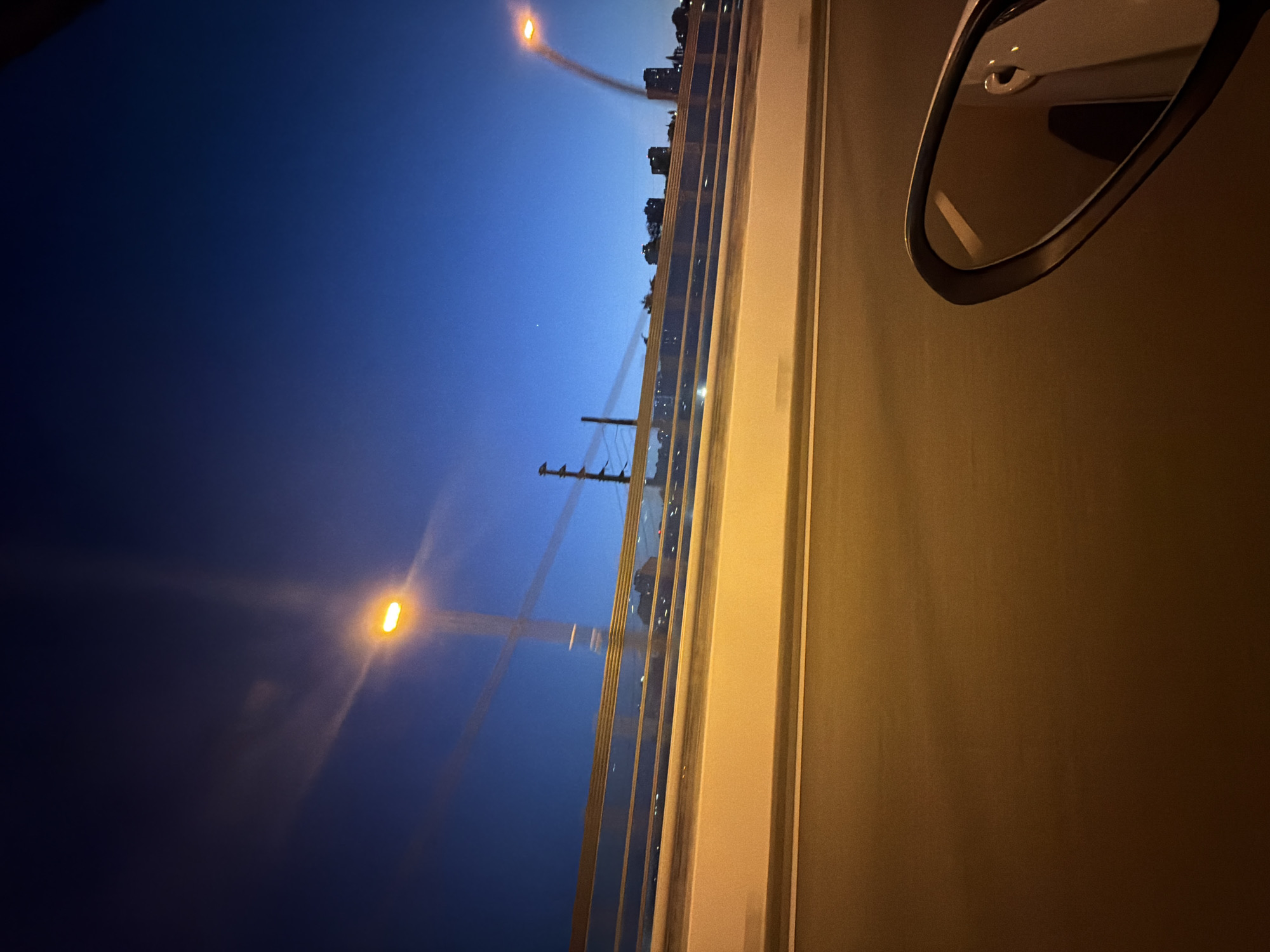 The image depicts a nighttime scene captured from a moving vehicle, likely on a bridge, overlooking a cityscape. The sky is a deep, clear blue, transitioning to a darker hue at the top. A bright orange streetlamp is visible in the upper left quadrant, its light streaking across the sky due to motion blur. Another similar streetlamp is visible in the upper right. Below the sky, a horizontal barrier, possibly a railing or guardrail, runs across the mid-section of the image. This barrier has a reflective, metallic surface and appears to be illuminated by the streetlights, casting streaks of light. Beyond the barrier, the silhouette of a city skyline with several buildings is discernible against the dark sky. The foreground consists of a road or highway, also showing signs of motion blur, with distinct lanes marked by lighter lines. In the lower right corner, a side-view mirror of the vehicle is visible, reflecting a portion of the car's interior and the dark sky. The overall impression is one of a nighttime drive through an urban environment, with the artificial lights of the city creating a dynamic and somewhat abstract visual experience. This scene could be representative of a location in Sochi, Russia, given the presence of urban infrastructure and a coastal city vibe implied by the distant lights.