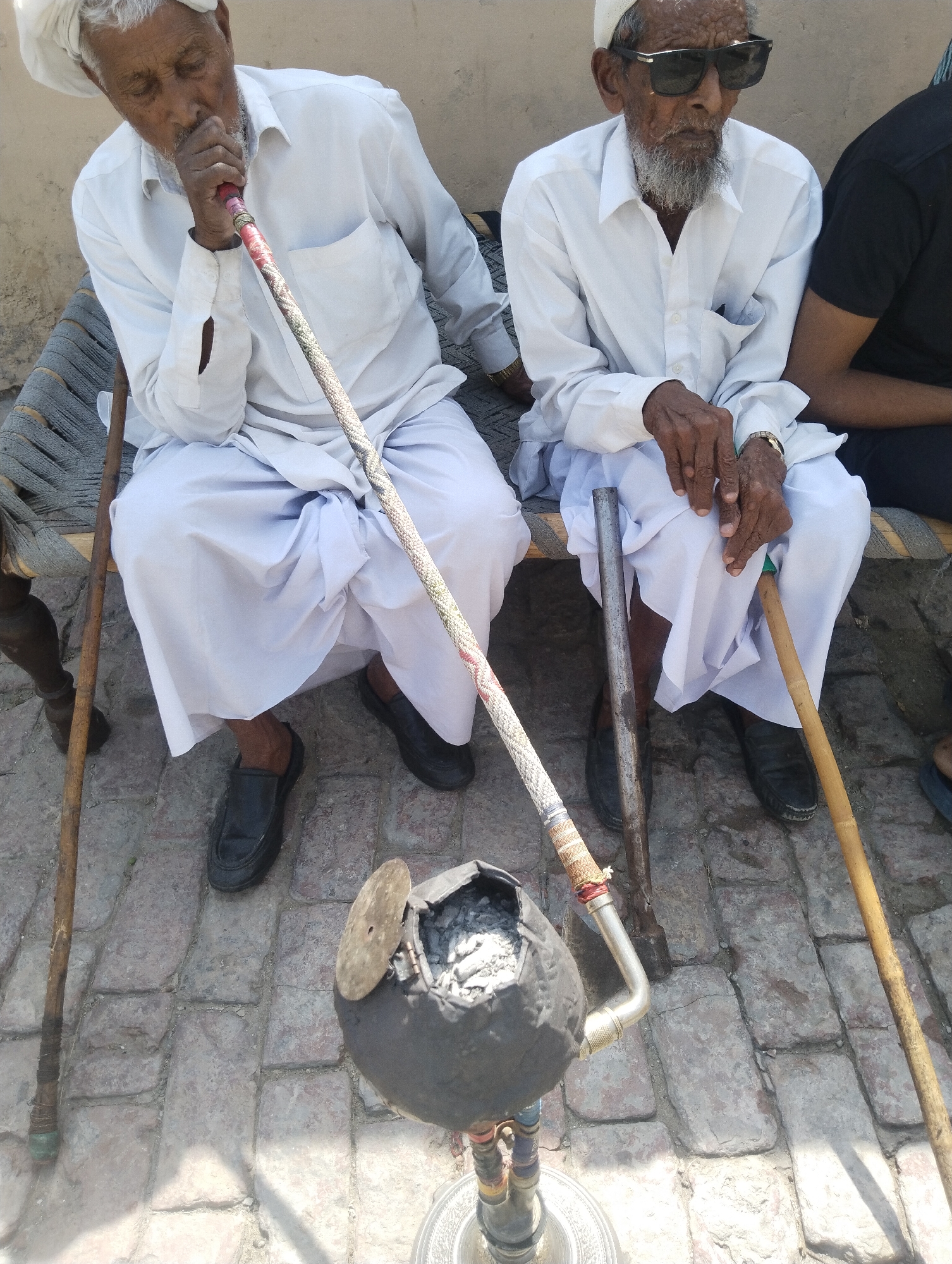 Two older men sit on a cot outdoors in Dera Chahal, Pakistan. The man on the left wears a white head covering, a white button-down shirt, and white pants, and he is smoking from a large hookah placed in front of them. The man on the right wears dark sunglasses, a white head covering, a white button-down shirt, and white pants. Each man has a walking stick resting beside him. A third person is partially visible on the right edge of the frame, wearing a black shirt. The setting is a paved area.
