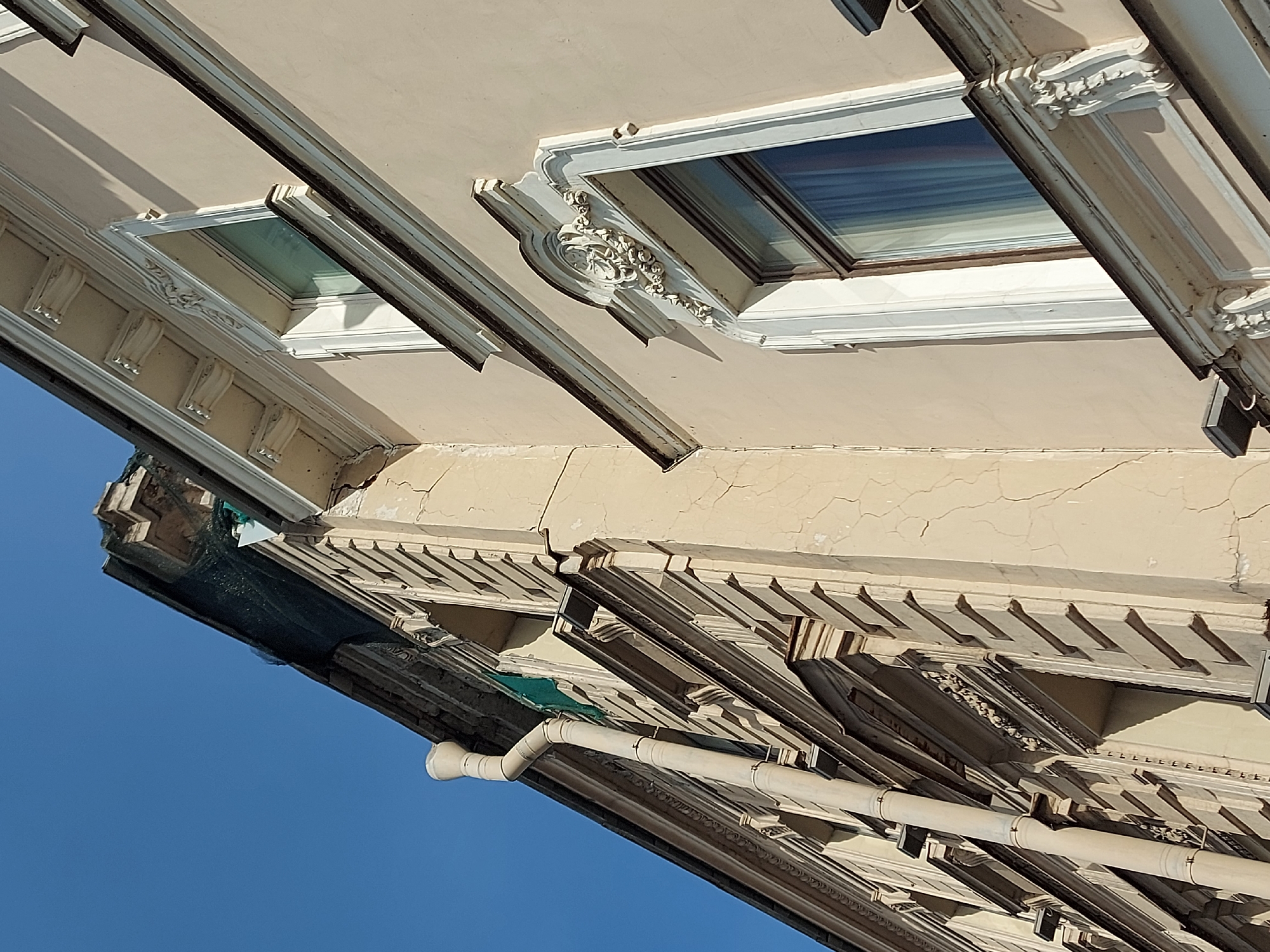 The image shows a low-angle view of an ornately decorated building facade against a clear blue sky. Architectural details include decorative moldings, window frames with ornate embellishments, and a rain gutter system attached to the building. The building's exterior appears to be aged, with visible cracks and imperfections. The architectural style and details suggest a location in a historic urban area, potentially in the city of Saint Petersburg, Russia.