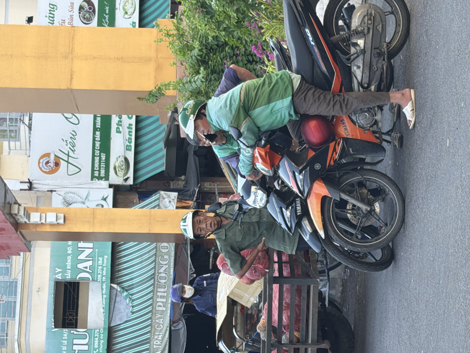 The image captures a street scene in Nha Trang, Vietnam, possibly near a market or food stall area. In the foreground, two men are seated on motorcycles. The man on the left, wearing a green uniform and helmet, has a weathered appearance and is looking towards the camera with a concerned expression, his right hand on his hip. He is standing next to a cart loaded with large, red, net-covered produce, possibly onions or potatoes. The man on the right, also wearing a green uniform and helmet, is talking on a mobile phone while leaning on his handlebars. His motorcycle is parked next to the other. Behind them, a woman wearing a blue uniform, mask, and helmet is visible, standing near the produce cart. Various signs and awnings are present in the background, suggesting commercial activity. The overall environment appears to be an outdoor urban setting, with buildings and greenery visible. The lighting suggests it is daytime, possibly late afternoon.