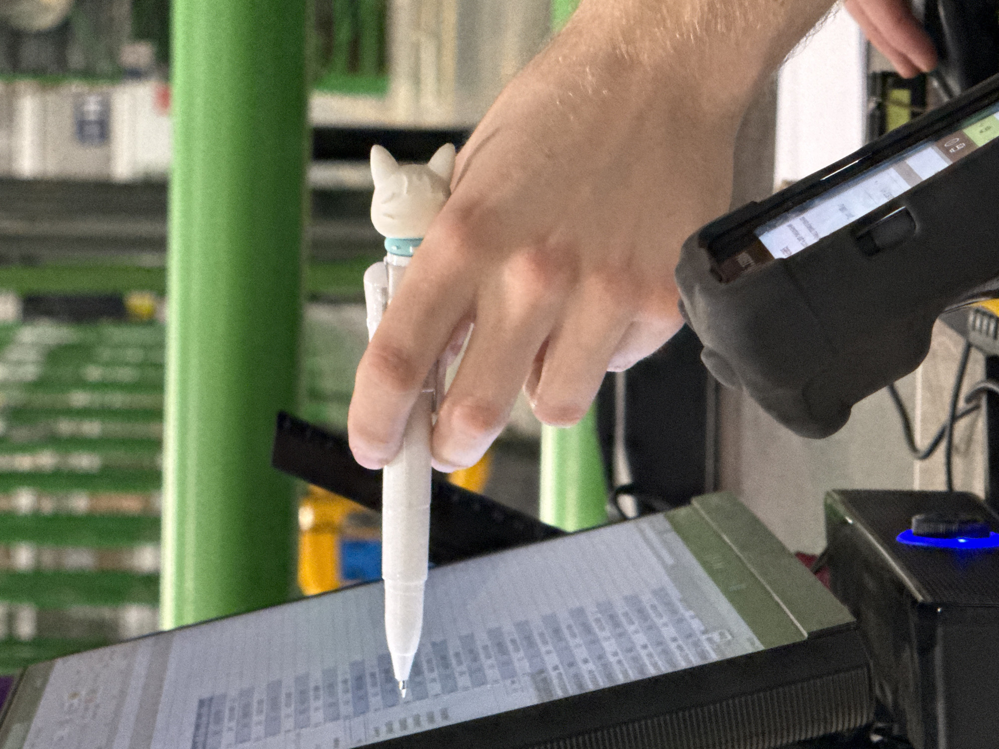 This close-up image shows a person's right hand using a white stylus with a small cat figurine to interact with a digital screen. The cat figurine, at the top of the stylus, is white with closed eyes and a light blue collar. The stylus's tip is positioned over a light-colored screen displaying rows and columns of text or data, possibly a spreadsheet or inventory list.
In the left portion of the frame, the same hand or another hand is holding a black, ruggedized handheld device, likely a mobile scanner or data terminal. Its screen is also visible, showing icons at the top (e.g., WiFi signal, battery indicator) and lines of text below, with one line possibly starting with "Smart...".
The setting appears to be an indoor work environment. The background is blurred, featuring prominent green vertical structures, possibly shelving or support beams, and hints of yellow and white objects. To the right of the main screen, a black rectangular device with a circular, illuminated blue button or dial is visible, connected by wires. The overall activity suggests a task involving data entry, inventory management, or similar operational work. No specific time of day or emotions are discernible. The provided location context is Lupanovo, Russia.