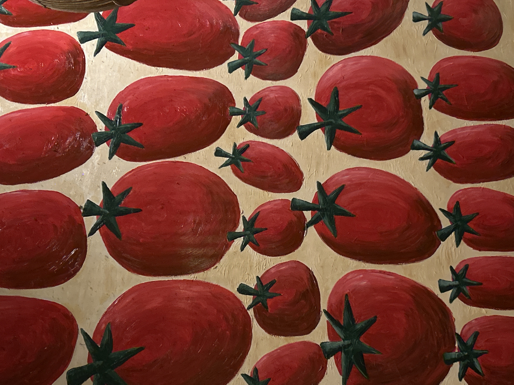 A high-angle shot shows a tightly packed arrangement of painted tomatoes against a textured, beige background. The tomatoes vary in size and shape, with most being oval or round. Their red color is applied with visible brushstrokes, creating a sense of depth and texture. Each tomato has a green star-shaped sepal. The painting is located in North Kuta, Indonesia.