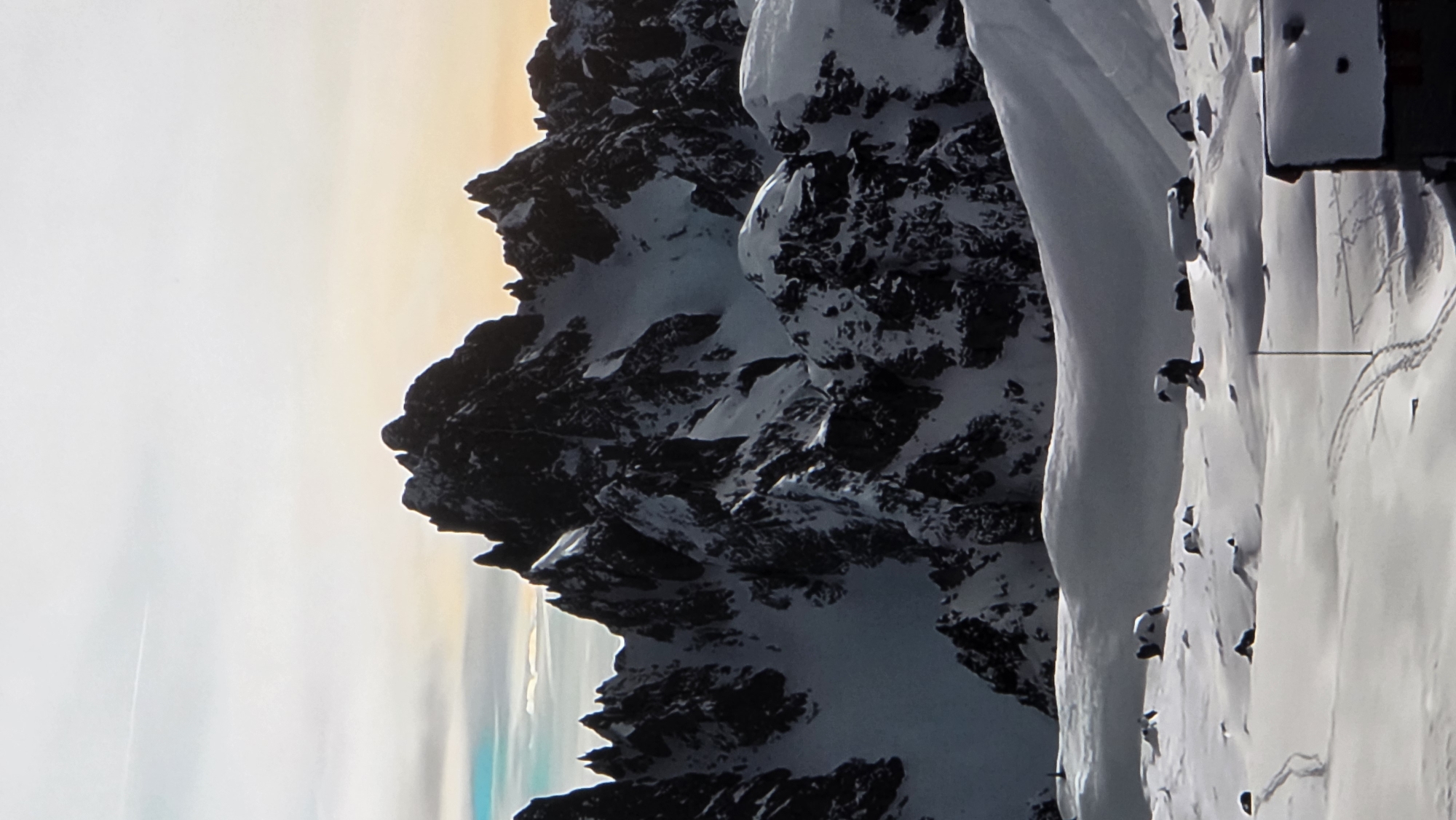 The image depicts a majestic mountain range covered in snow under a sky with wispy clouds and a hint of sunset colors. The foreground features a vast expanse of untouched snow, interspersed with dark rocks peeking through the white blanket. In the far distance, a small, dark structure with a snow-covered roof is visible on the right side of the frame, suggesting a human presence in this remote, mountainous environment. There are also ski tracks visible in the snow in the foreground, indicating activity. The scene is one of serene, natural beauty and rugged wilderness. This image is not located in the city of Batumi, Georgia; it depicts a snow-covered mountain landscape.