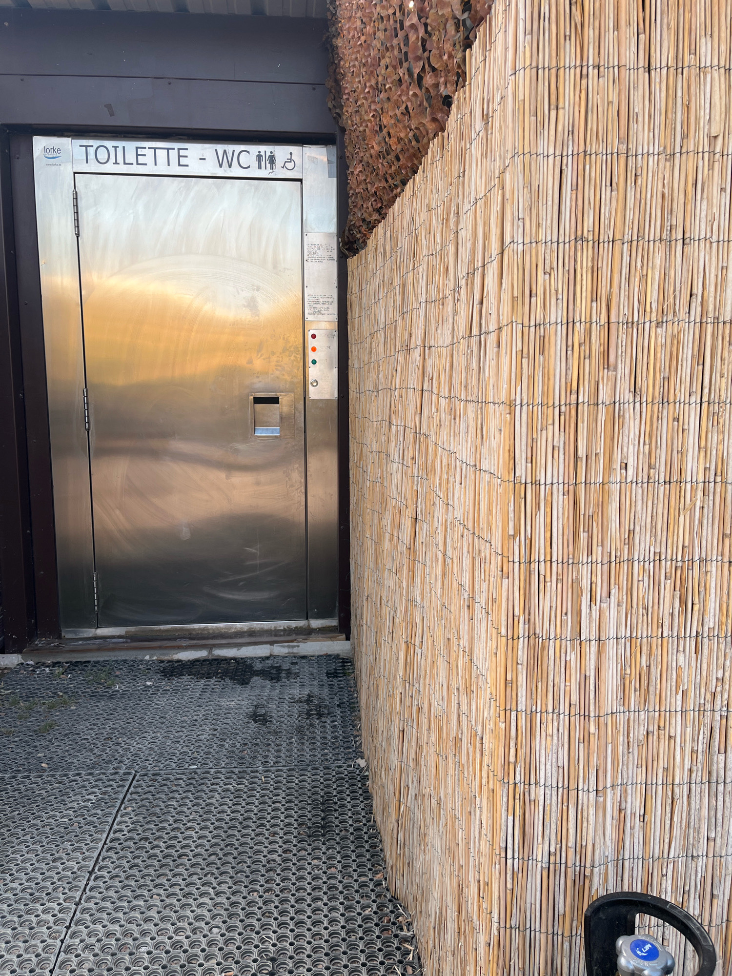 The image shows the exterior of a public restroom facility located in Saint-Just-Saint-Rambert, France. The entrance features a brushed metal door with a rectangular push-button mechanism and signage indicating "TOILETTES - WC" with universal accessibility symbols for men, women, and individuals with disabilities. To the right of the door, a notice board displays a series of colored lights (red, orange, green) and what appears to be a coin or card reader. The restroom structure is bordered by a fence constructed from vertical bamboo poles, bound together with dark wire, and a camouflage-like netting is visible at the top of the structure. The ground in front of the restroom is covered with a dark, perforated rubber matting system. A black propane tank with a blue and silver valve is partially visible on the right side of the frame. The overall scene suggests an outdoor, public amenity.