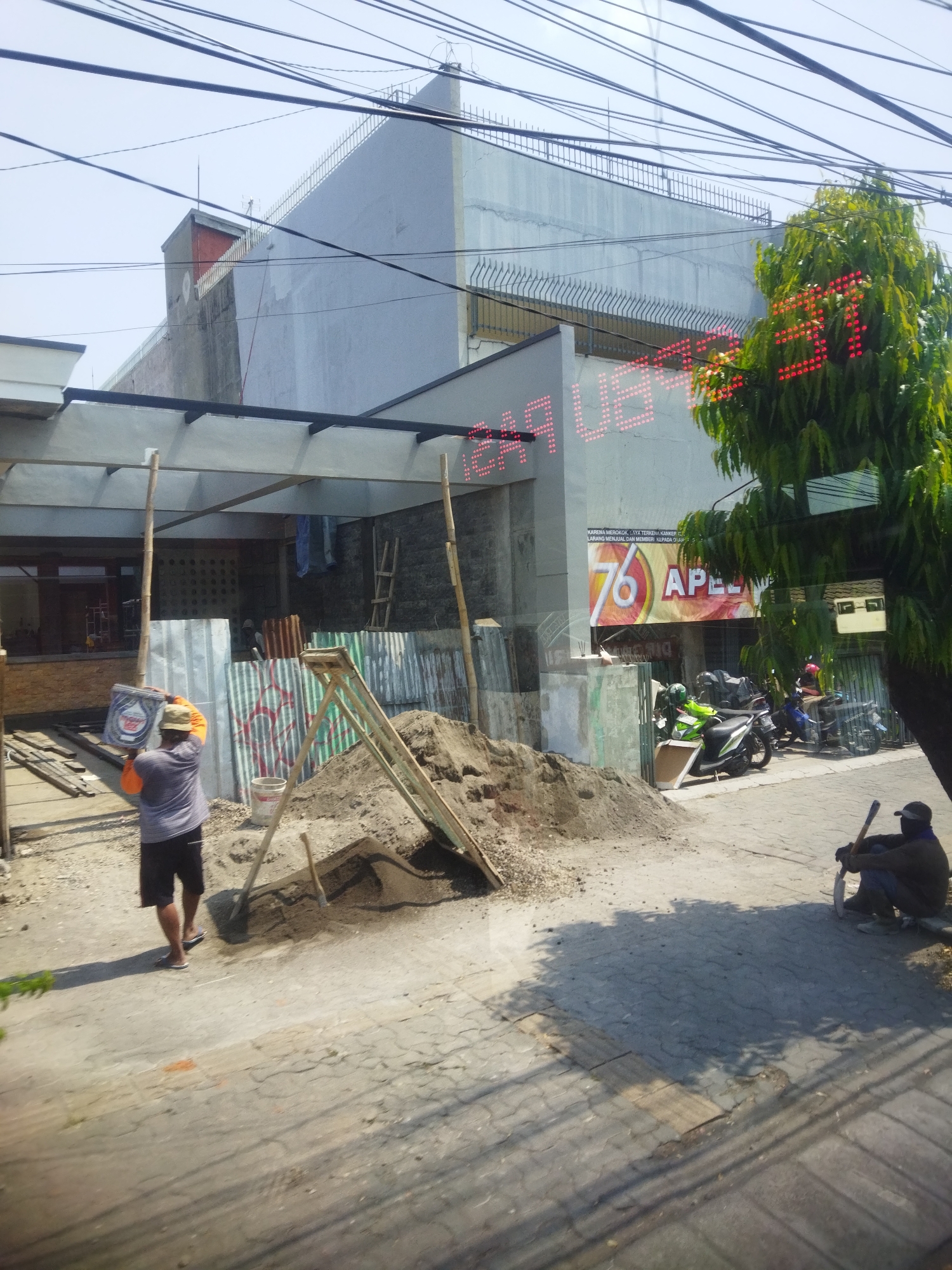 A construction site is depicted in front of a building with a large "76 APEL" banner. A man in a gray shirt and black shorts carries a bucket, while another man in a dark shirt and blue jeans sits on a curb, holding a shovel. Several motorcycles are parked near the building. The scene is set on a sunny day with a clear sky. The location is identified as Surakarta, Indonesia, based on the "76 APEL" advertisement which is a popular gas station chain in Indonesia.