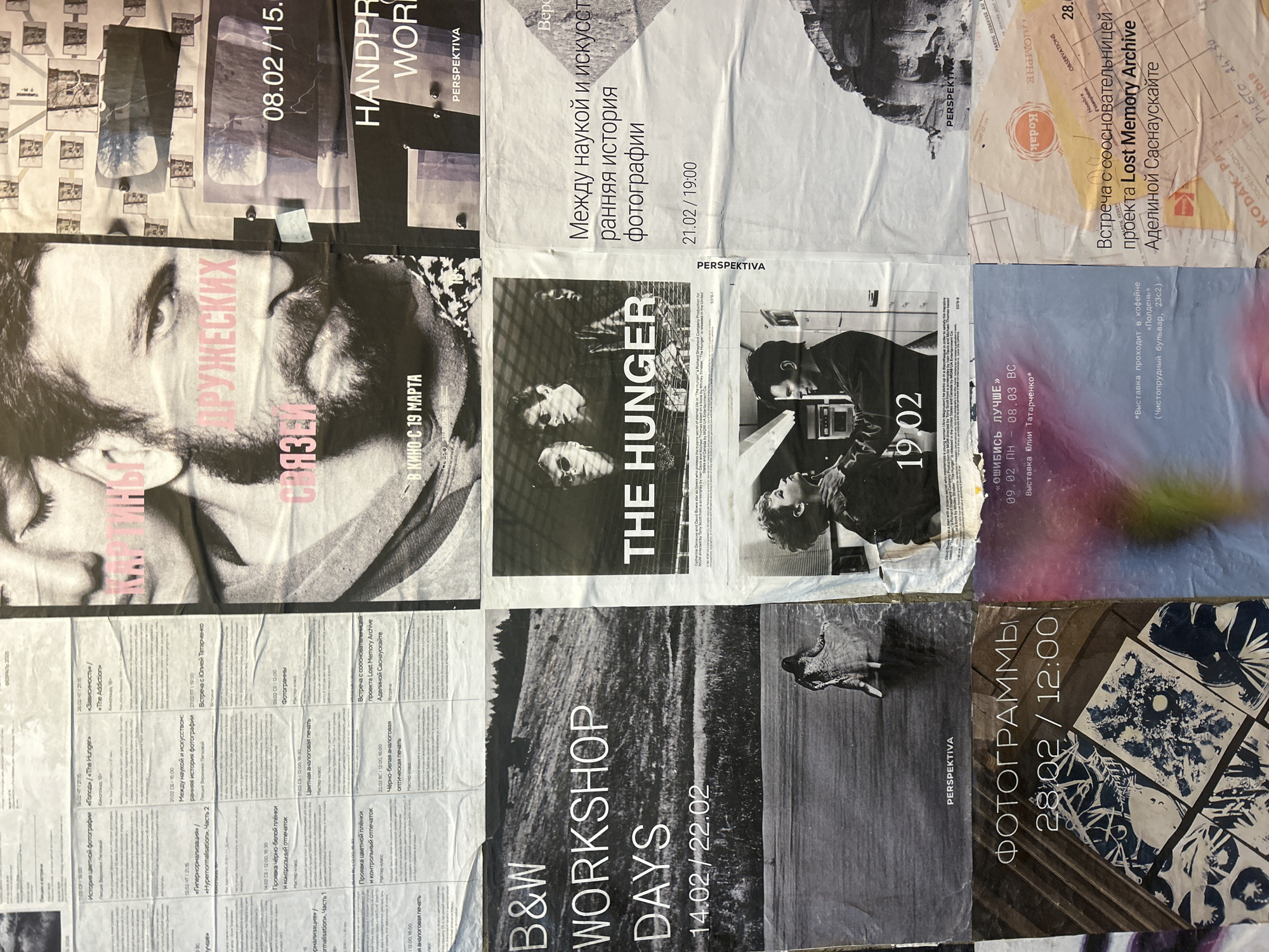This is a close-up image of a wall covered with posters, likely in Moscow, Russia, given the Cyrillic text and specific locations mentioned. The posters promote various events and workshops related to photography, film, and art. The topics covered include the history of photography, black and white and color analog printing, and screenings of films like "The Hunger." Some posters highlight specific dates and times for these events, such as a workshop on February 14-22, 2022, and film screenings starting March 19th. There is also a poster advertising an exhibition by Yulia Tatarchenko at the Polden cafe on Chistoprudny Boulevard. One poster mentions a meeting related to the "Lost Memory Archive" project. Overall, the scene appears to be a bulletin board or wall showcasing announcements for artistic and cultural events.