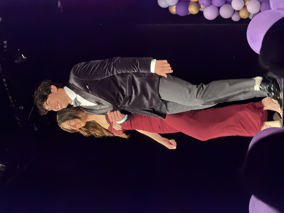 This image captures a male and a female on what appears to be a stage in Palos Verdes Estates, United States. The male, positioned slightly forward on the right, exhibits dark, curly hair and a smiling expression directed towards the right. He is attired in a dark, possibly velvet, suit jacket over a white collared shirt, a dark vest, a grey bowtie, grey trousers, white socks, and shiny black dress shoes. His right hand is partially clenched. The female, positioned slightly behind and to the left of the male, has long brown hair and a matching smiling expression, also directed towards the right. She wears a strapless, ruched, floor-length deep red gown and light-colored open-toed high heels. Both individuals appear to be in motion, walking from left to right across the frame. The background is predominantly dark, suggesting a stage setting, with some overhead stage lighting fixtures visible at the top. To the far right, a vertical arrangement of balloons in shades of purple, light purple, and gold serves as decoration. In the immediate foreground, at the bottom, blurred purple objects, likely balloons, are partially visible, indicating a perspective from the audience level.