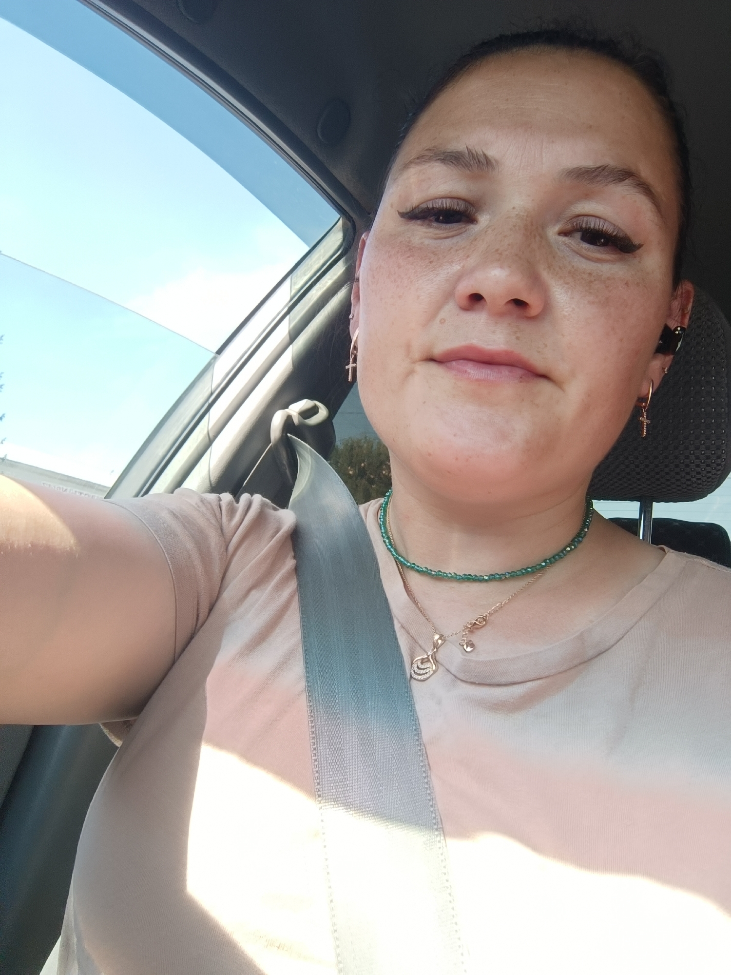A woman is seated in the passenger seat of a car, looking directly at the camera. She has freckles on her face and is wearing a tan t-shirt. A gray seatbelt is visible across her chest. She is also wearing a green beaded necklace with a pendant and hoop earrings, one of which has a small cross. She has a black earbud in her right ear. Sunlight streams in from the side window, illuminating the car's interior and casting shadows on the woman's arm. The sky outside is blue. The scene appears to be in the city of Chaikovsky, Russia.