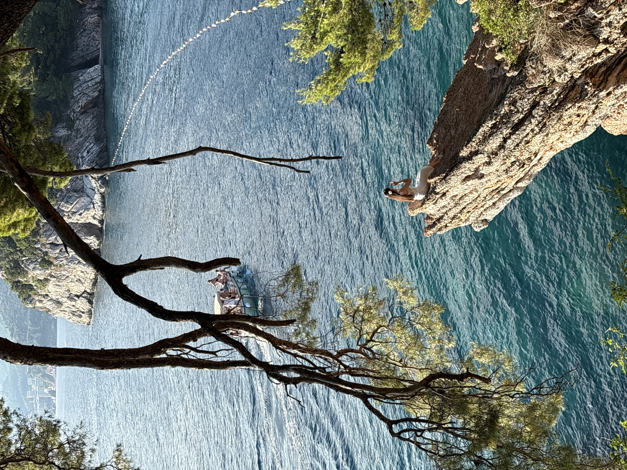A woman sits on a rocky outcrop overlooking the Adriatic Sea in Sveti Stefan, Montenegro. She is wearing a white dress and has her hair pulled back. She is holding a phone to her ear and appears to be taking a photo. The sea is a vibrant turquoise color, and a small boat with several people on board is visible in the distance. The coastline is rugged and mountainous, with lush green trees covering the slopes. The image is framed by the branches of a large pine tree.