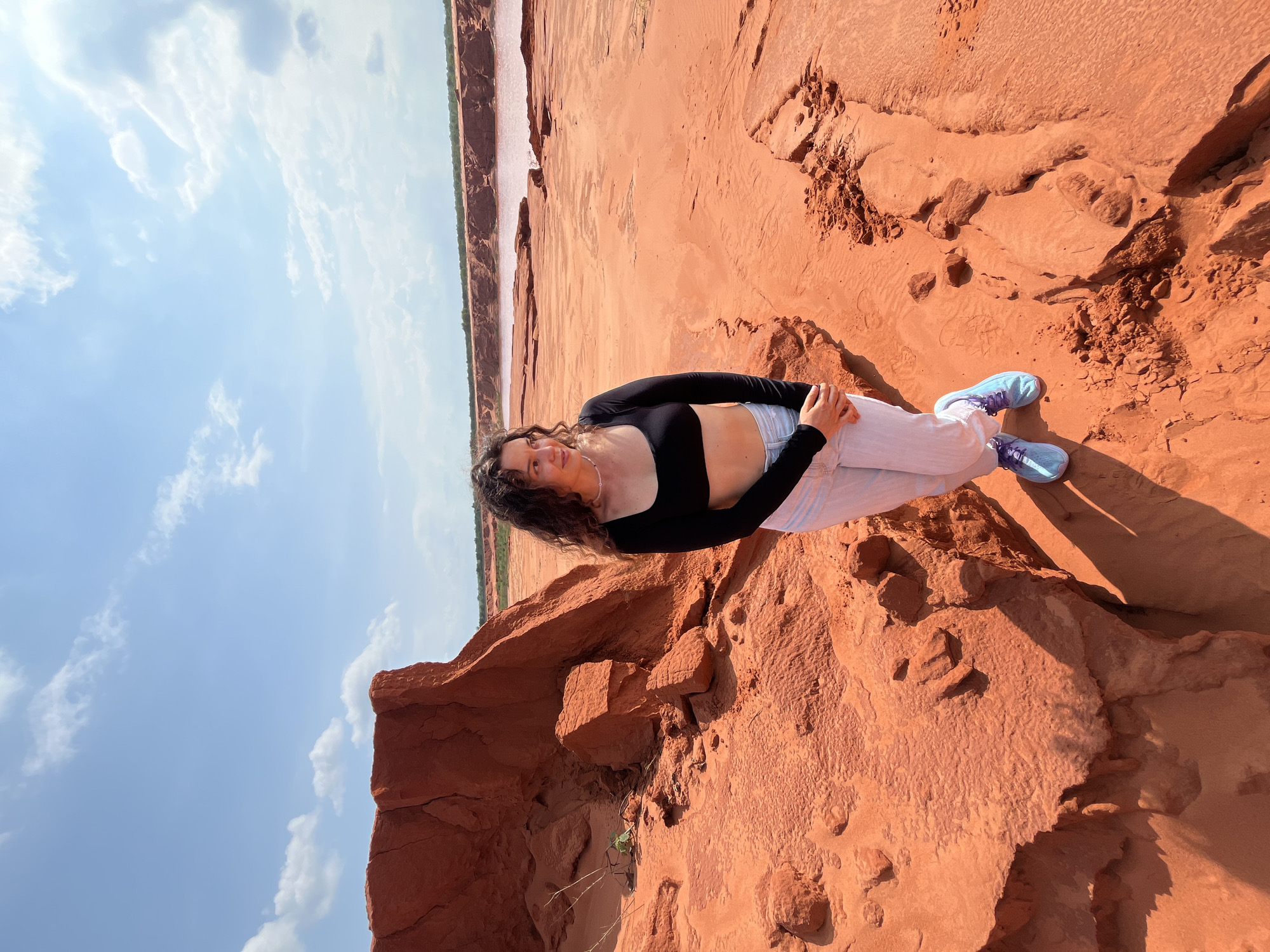 In Mui Ne, Vietnam, a young woman with curly brown hair stands in a desert-like landscape. She is wearing a black long-sleeved crop top, white pants, and light blue sneakers. The ground is covered in red sand and rock formations. In the background, a body of water with a pinkish hue stretches towards the horizon under a bright blue sky with scattered clouds. The scene captures the unique geological features of the area.