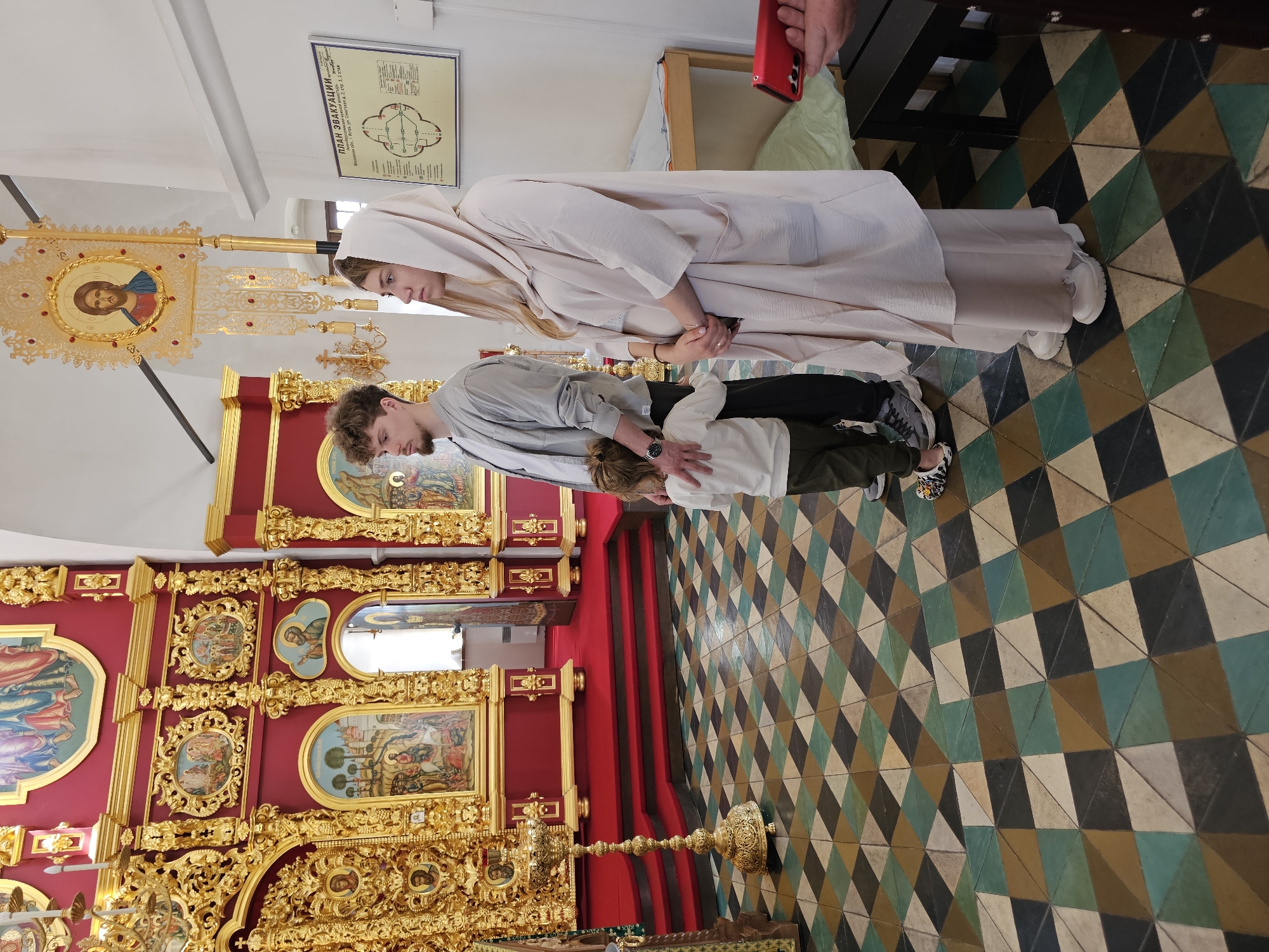 The image captures a scene inside what appears to be an Orthodox church in Istra, Russia, as suggested by the opulent, gold-detailed altar with traditional iconography. A young woman dressed in a long, cream-colored robe and head covering is standing beside a young man in a grey shirt. A child standing near the man is being held by the man. The three are positioned on the red carpeted area in front of the altar. The floor is covered in geometric-patterned tiles. In the upper part of the image, a plan of evacuation sign is visible.