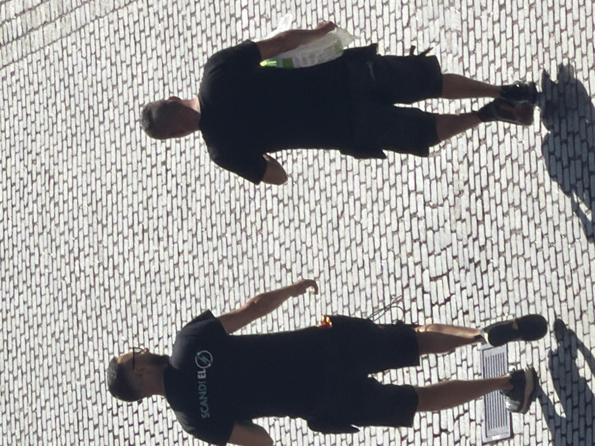 Two individuals, one wearing a black t-shirt with "SCANDI EL" printed on it and dark shorts, and the other wearing a black t-shirt and black shorts, are walking away from the camera on a brick-paved surface. The individual on the left has glasses and a tool belt attached to their shorts. The individual on the right is carrying a white plastic bag in their right hand. The scene appears to be an outdoor setting, likely a street or plaza, with the brick paving suggesting an urban environment in Malmö, Sweden. The lighting indicates it is daytime, with shadows cast on the ground by the individuals and the paving stones.