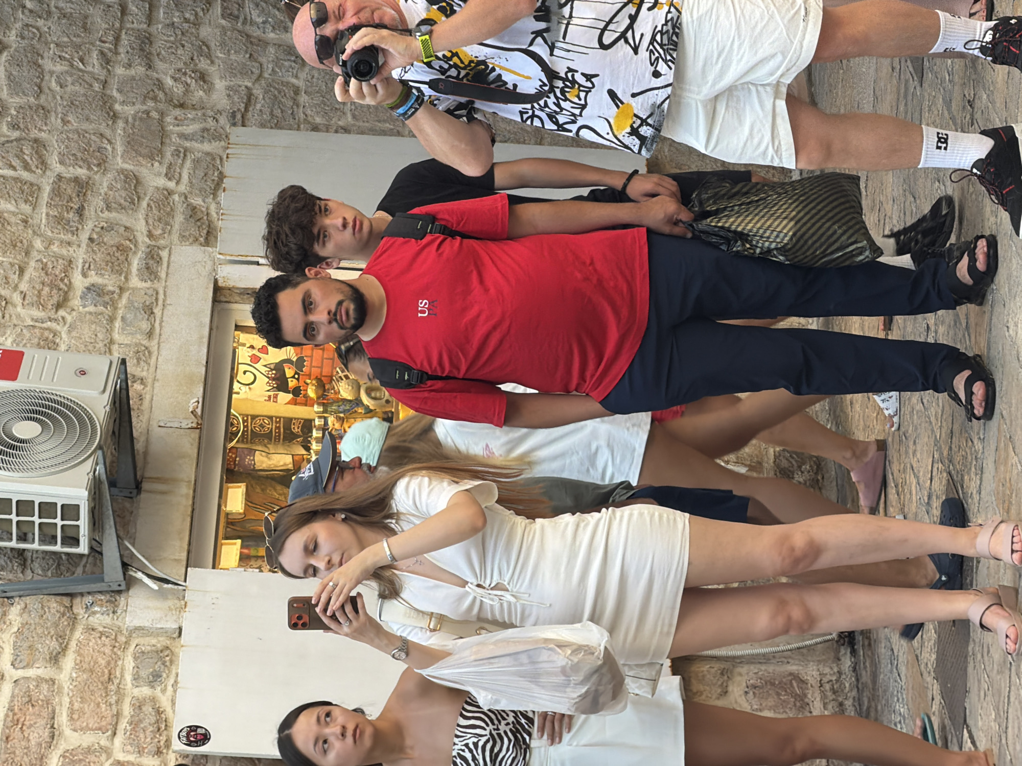 In Budva, Montenegro, a group of tourists is gathered outside a shop, possibly on a street. The architecture features a stone wall and a white storefront with an air conditioning unit mounted above.
In the foreground, a woman in a white dress holds a smartphone, appearing to take a photograph. Next to her, a young woman in a zebra-print top and white skirt looks away from the camera. To the right, a man in a red t-shirt and dark pants holds a black plastic bag. Behind him, a younger man in a black t-shirt looks towards the front. Further to the right, a man wearing sunglasses and a white shirt with a black and yellow graphic pattern holds a camera up to his eye, taking a picture. He is wearing white shorts. The ground is paved with cobblestones.