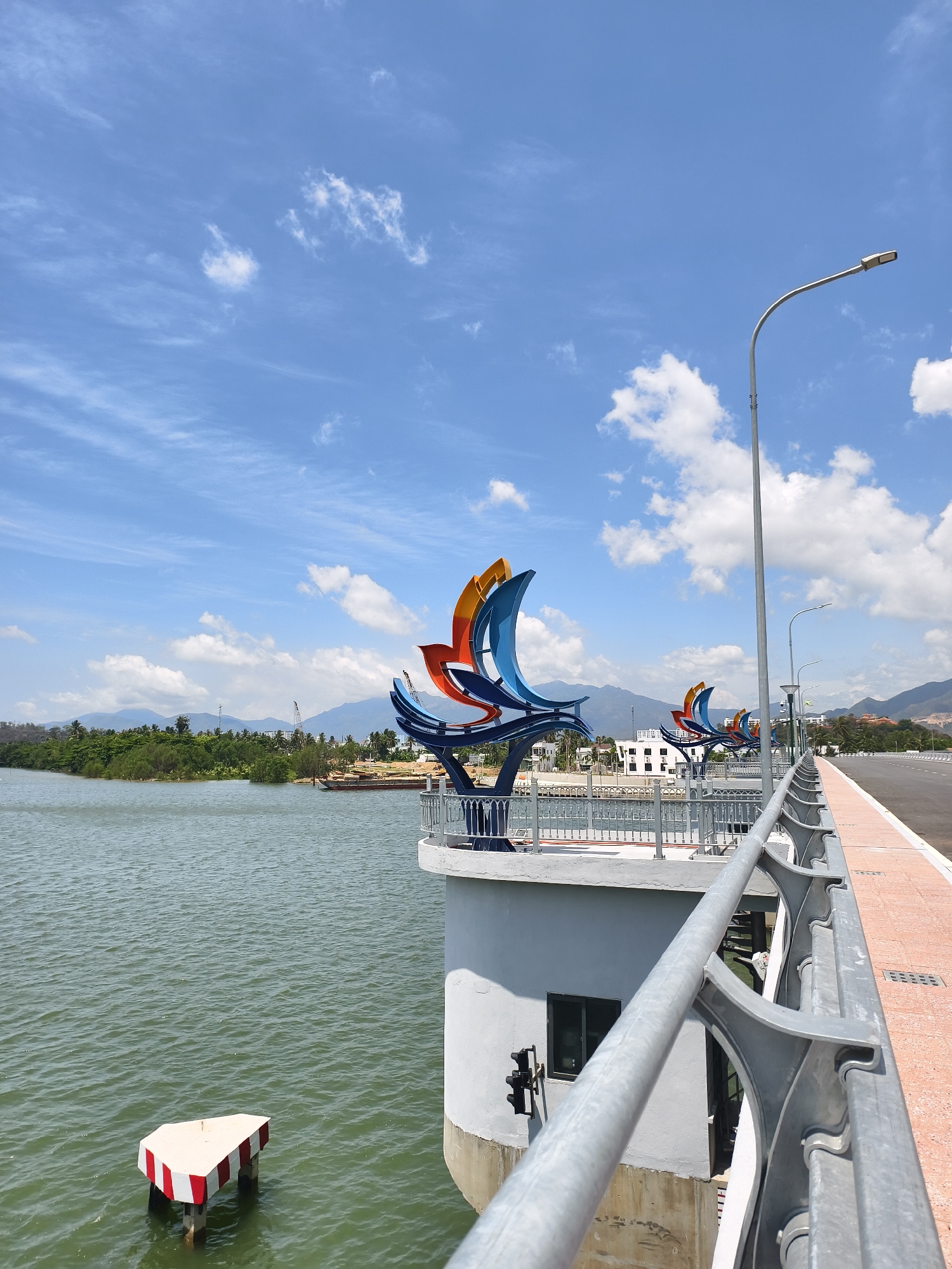 The image captures a bridge in Tay Nha Trang Ward, Vietnam. The bridge has a pedestrian walkway lined with grey metal railings and features large, colorful abstract sculptures resembling stylized sails or lotus petals in hues of blue, red, and orange. These sculptures are mounted on the edge of the bridge, above gray concrete supports. In the background, mountains rise, and there's a view of a body of water with a small, red-and-white striped navigational marker. The sky is blue with scattered white clouds, and the scene is bathed in sunlight. A street lamp post stands tall beside the bridge. The road surface is a reddish-brown color.