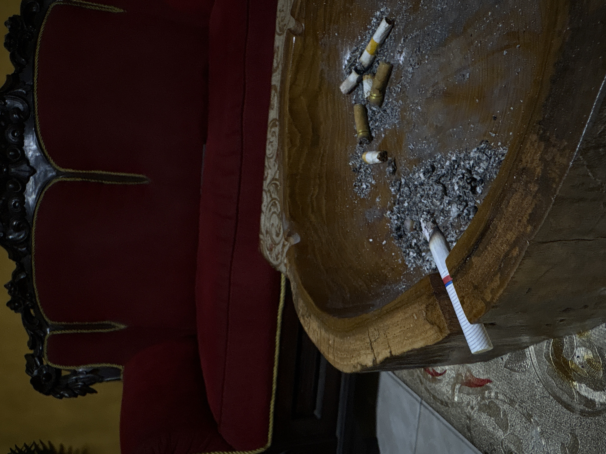 The image shows an interior scene in Kalijaten, Indonesia. A red, ornate armchair is positioned next to a large, wooden ashtray filled with cigarette butts and ash. One unsmoked cigarette rests on the edge of the ashtray. The ashtray appears to be carved from a single piece of wood. A patterned rug is visible beneath part of the ashtray. The scene appears indoors.