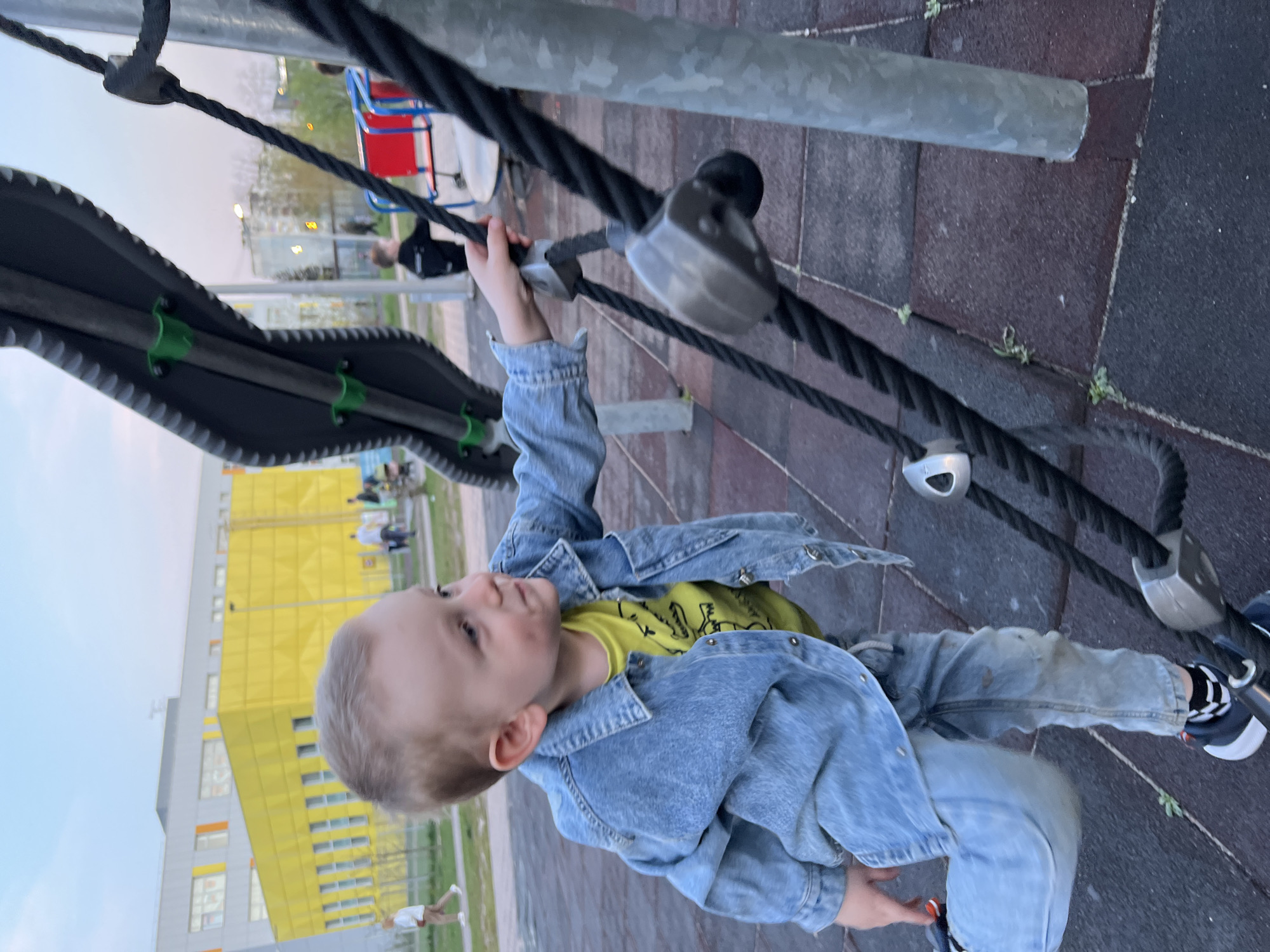 A young boy wearing a blue denim jacket over a yellow shirt and jeans plays on playground equipment made of metal poles, black ropes, and gray attachments. The boy is holding onto one of the ropes with his right hand. The playground has a red and white structure visible in the background. A yellow building with multiple windows is visible in the background. The scene is located in the city of Mytishchi, Russia.