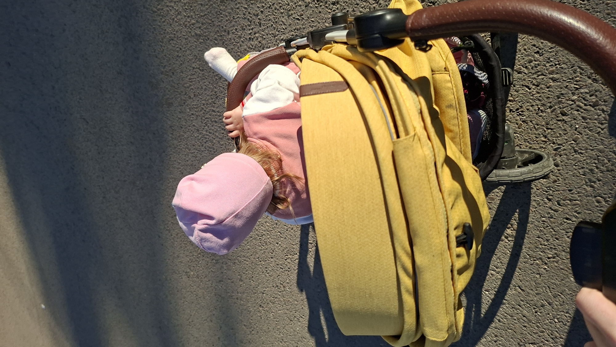 A high-angle, close-up photograph depicts a baby in a stroller, positioned next to a yellow backpack. The baby wears a pink hat and a pink and white outfit. One small hand is visible, as is the back of the baby’s head showing some reddish-brown hair. The stroller is on a paved surface. The location in the city of Chaikovsky, Russia, cannot be determined from this image.