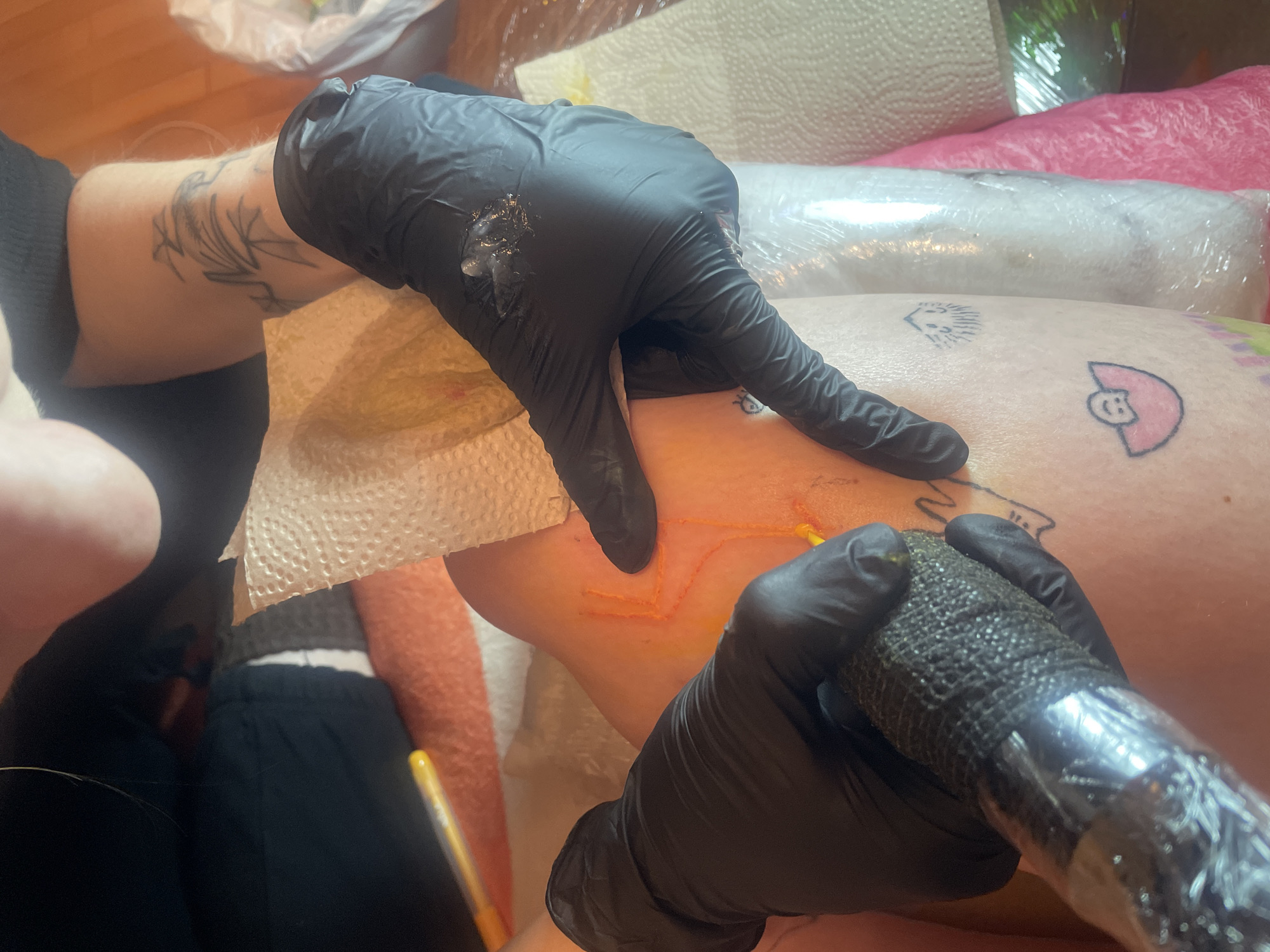 A person is getting a tattoo on their leg. The tattoo is outlined in orange and appears to be a geometric design. The tattoo artist is wearing black gloves and using a tattoo machine. The person's leg already has other small tattoos, including a cartoon character and a hedgehog. The scene is taking place indoors, likely in a tattoo parlor or studio. The location is in Vracar Urban Municipality, Serbia.