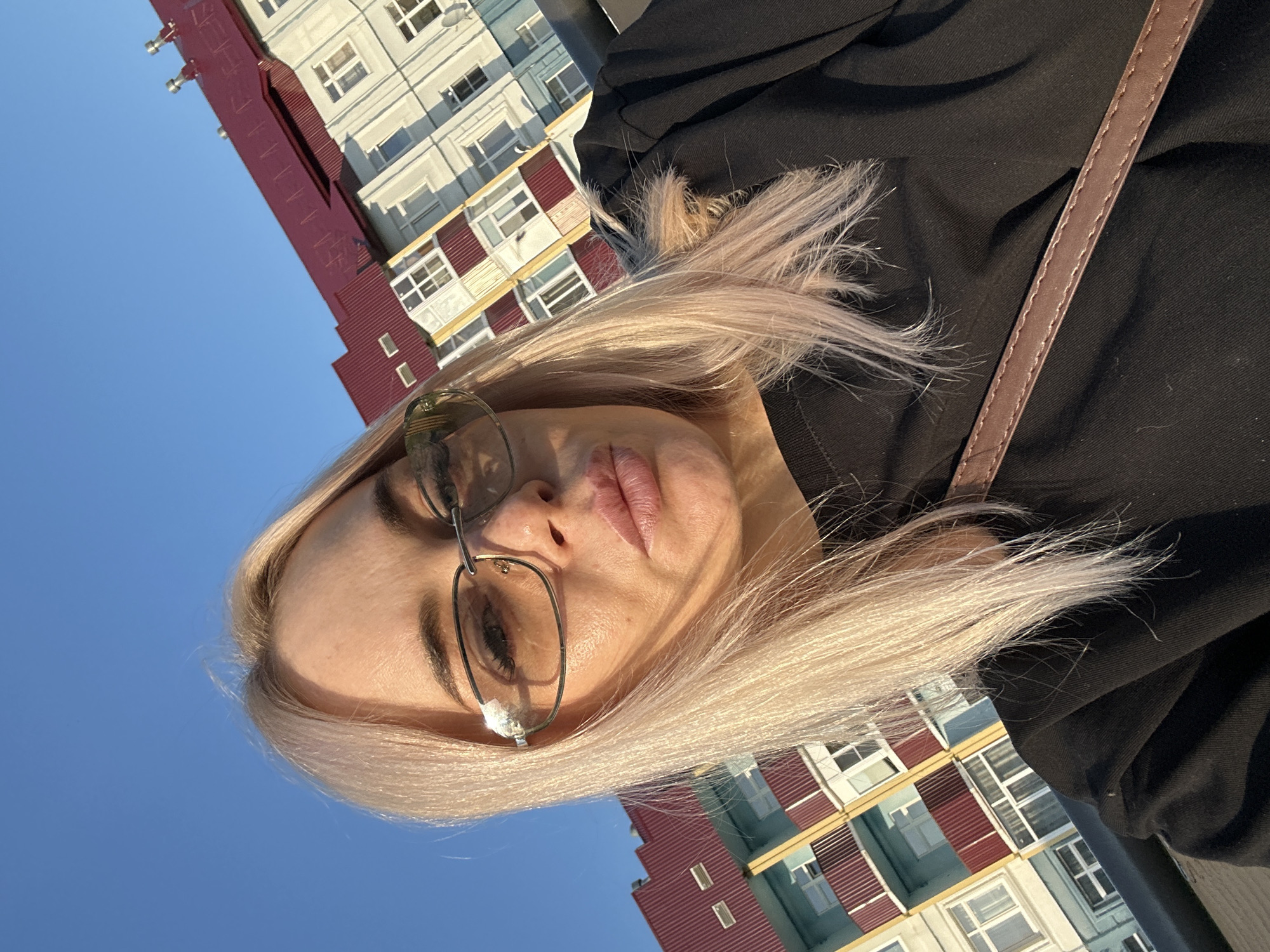 A woman with blonde hair and sunglasses takes a selfie outdoors with a building in the background. The building has a red roof and light-colored walls with balconies. The sky is clear and blue. The woman is wearing a black top and a brown strap from a bag is visible.