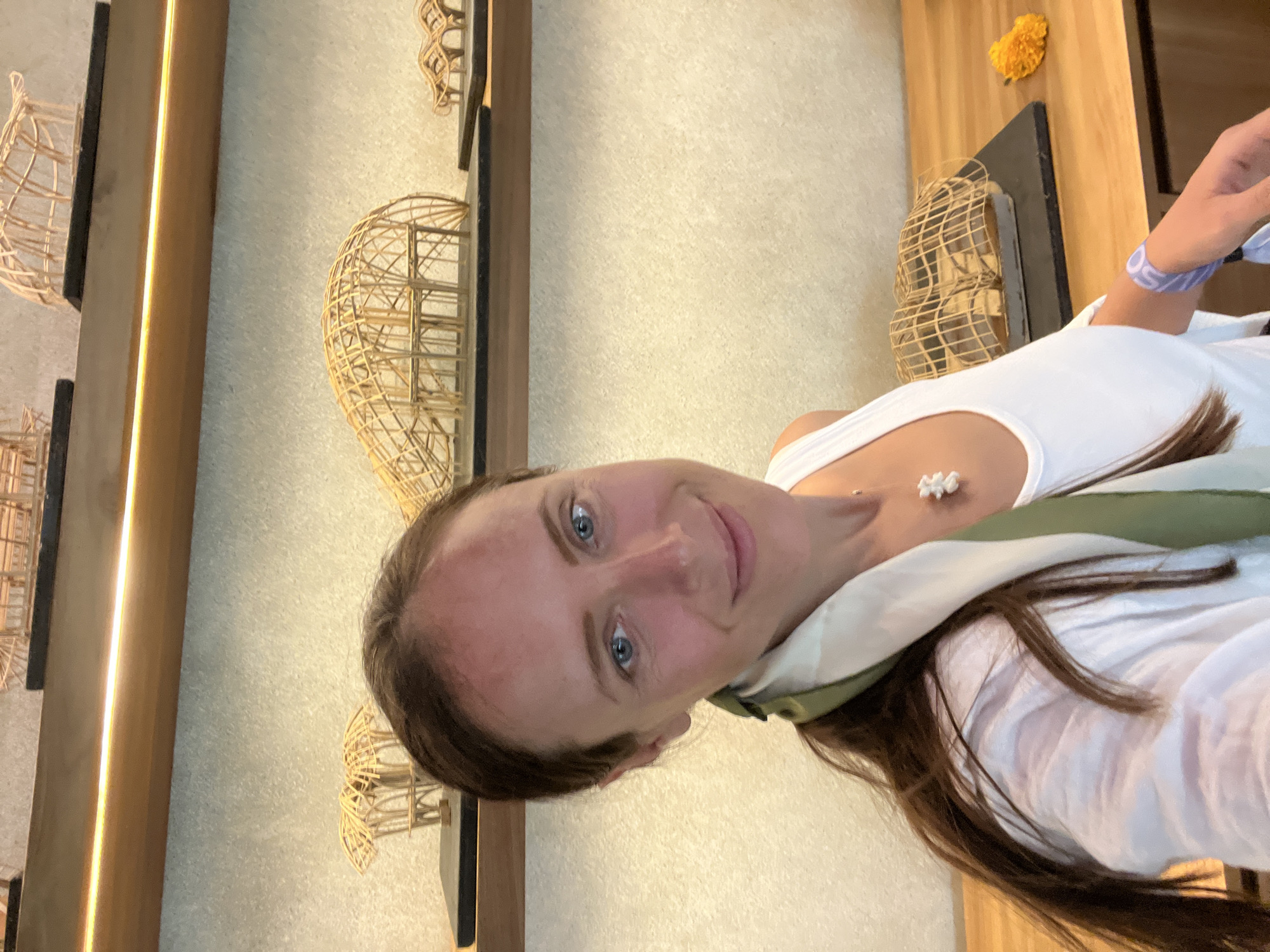 A female individual, appearing to be in her mid-20s to late 30s, is captured in a self-portrait, smiling directly at the camera. Her long, dark brown hair is pulled back, and she has blue eyes. A slight flush or redness is visible on her forehead. She is wearing a white tank top underneath a translucent white long-sleeved shirt, which is open at the front. A green and off-white patterned scarf is draped around her neck and shoulder. A small, white, textured necklace, possibly a cluster of shells or carved elements, hangs from a thin chain around her neck. A purple and white wristband is visible on her right wrist, while her right hand, holding the camera, is partially visible in the bottom right corner.
The setting is an indoor exhibition space, resembling an architectural design studio or gallery, located in the Ubud District, Indonesia. The background features light beige, textured walls. Two dark wooden shelves are mounted horizontally, with the upper shelf incorporating a warm LED strip light that illuminates the shelf below. Both shelves display multiple intricate architectural models, constructed from light-colored wood or bamboo sticks. These models depict various organic, curvilinear structures, such as domes, pods, and complex frameworks, each resting on a flat, dark rectangular base. On a light wooden counter in the foreground, below the lower shelf, another large, enclosed model is visible behind the subject's right arm. A single bright yellow marigold flower rests on this wooden counter to the right of the model.