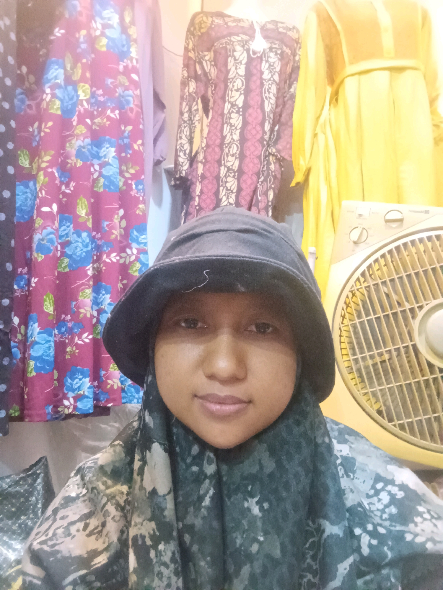 A young woman in a black bucket hat and a patterned hijab poses for a selfie indoors. She is positioned in the foreground, with a bright yellow fan to her right. Behind her, colorful fabrics with floral and geometric patterns are visible, hanging on what appears to be a clothing rack or display. The background suggests a market or retail setting. The lighting is somewhat diffused, and the overall scene is casual and personal. The location is identified as Sumuragung, Indonesia.