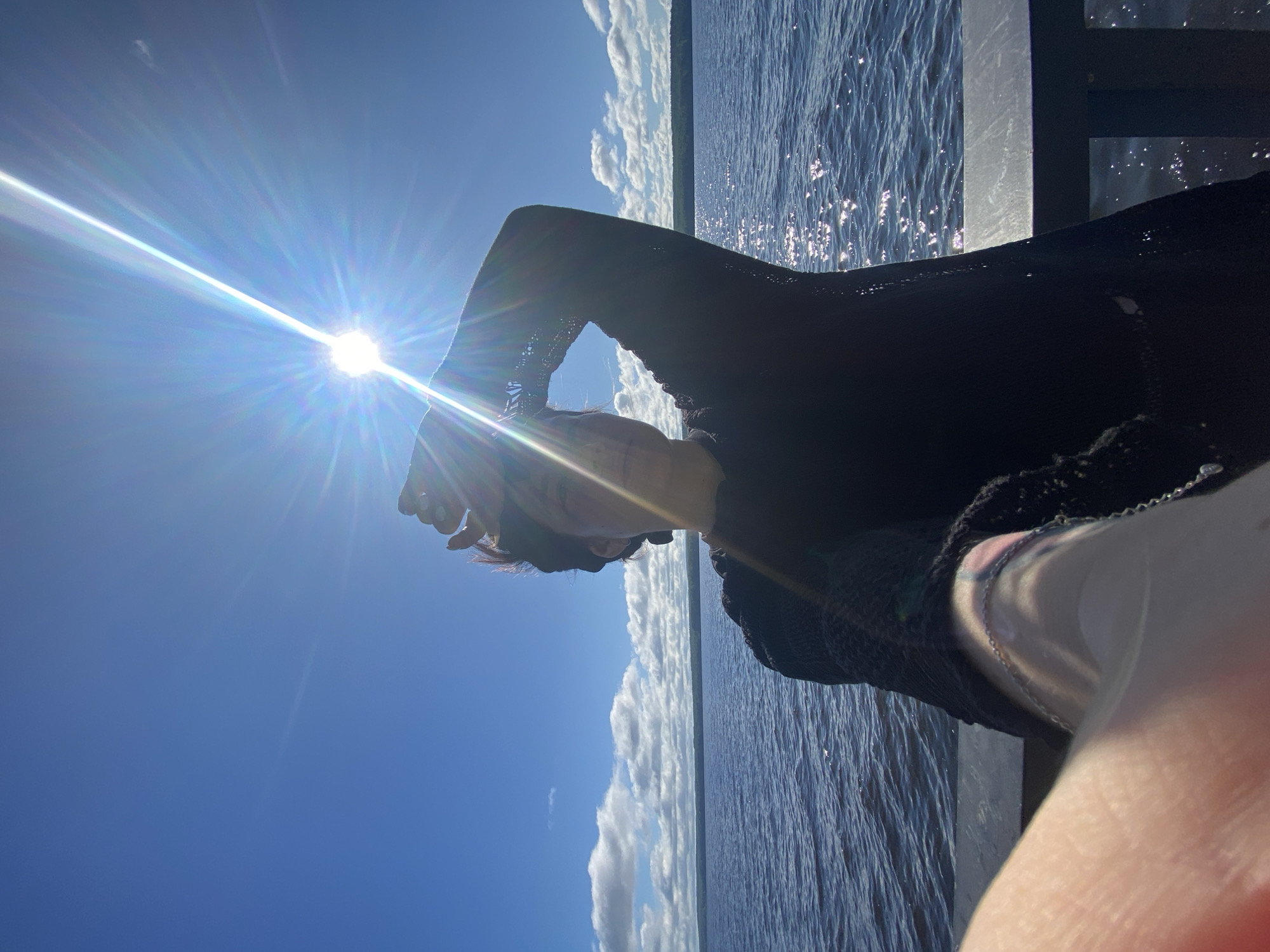 A person is standing on a pier, their face partially obscured by their hand raised to shield their eyes from the bright sun. The sun is positioned in the upper center of the frame, creating a strong lens flare that radiates outward. The person is wearing a dark top. Behind them, a large body of water stretches to the horizon, with a tree line visible in the distance under a blue sky dotted with white clouds. The water is choppy and reflects the sunlight. The location is identified as the Kama River in the city of Chaikovsky, Russia.
