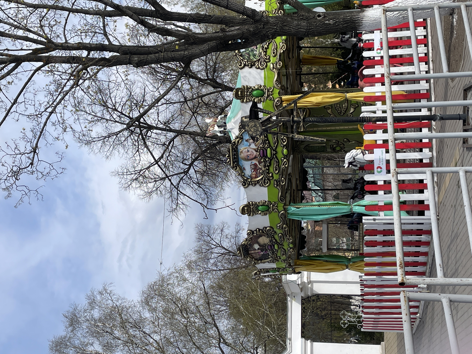 The image depicts a colorful carousel in what is likely a park or public space in Urussu, Russia. The carousel is decorated with images of Disney characters. It is enclosed by a red and white striped fence. The scene is partially obscured by bare trees. The sky is overcast. Several individuals are standing near the carousel.