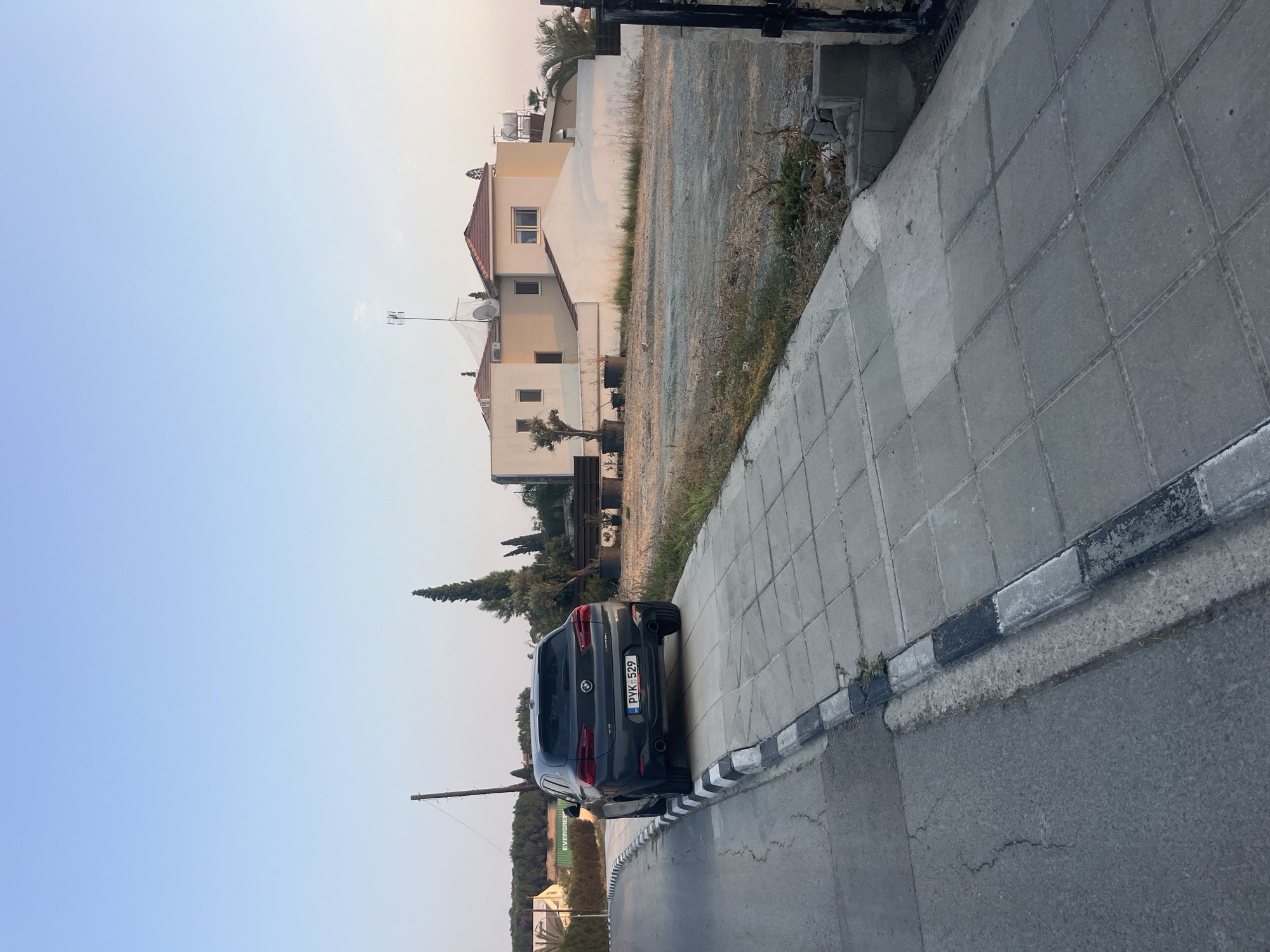 In Parekklisia Community, Cyprus, a dark grey BMW X2 is parked on a paved driveway that slopes upwards from the street. The car faces away from the camera, its rear end prominently displayed. The license plate reads "PYK 529". To the left of the car, a sidewalk with alternating black and white striped curbing runs alongside a paved road. In the background, a tan and cream-colored house with a red-tiled roof is visible, situated on a lot with sparse vegetation and dry grass. Tall, slender trees, possibly cypress, are scattered around the property. A utility pole stands near the road. The sky is a clear, pale blue, suggesting it is either early morning or late afternoon. The scene appears quiet and residential.