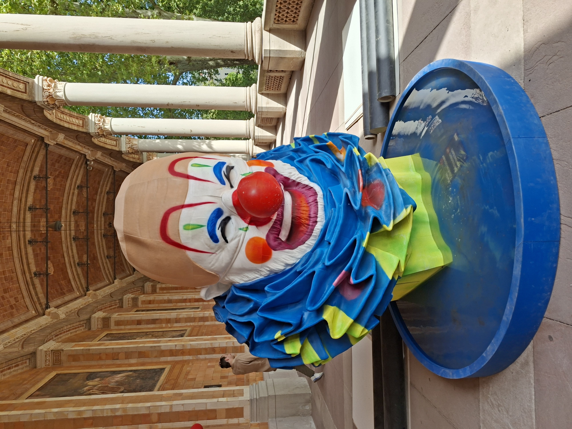 This image shows a large clown head sculpture located in the Kurhaus colonnade in Baden-Baden, Germany. The sculpture is positioned on a blue circular platform filled with water. The clown head is brightly colored with a red nose, painted blue and red facial features, and a large blue and yellow ruffled collar. Behind the sculpture, the architectural features of the Kurhaus are visible, including arched ceilings, tiled walls, and ornate columns. To the right, large trees with green foliage are seen, indicating an outdoor setting. In the background, a person wearing a brown shirt and dark pants is walking away from the camera. Rolls of dark material are placed on the ground near the sculpture. The lighting suggests it is daytime.