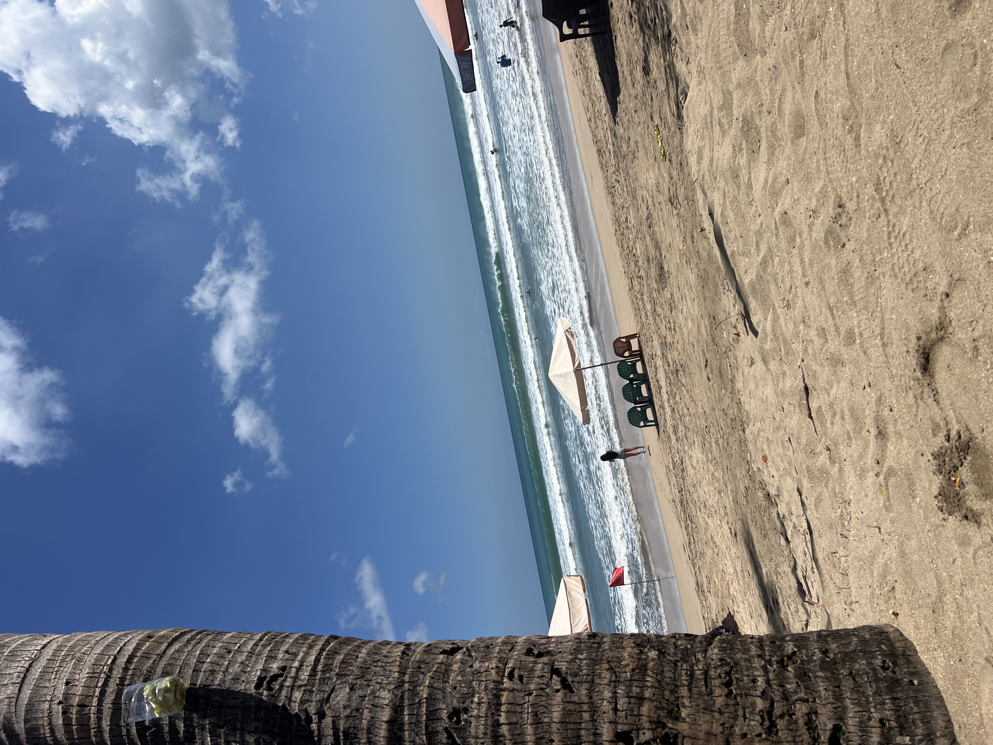 A bright, sunny day at Kuta Beach, Bali, Indonesia. The foreground is dominated by the textured bark of a palm tree on the left. The sandy beach stretches towards the ocean, where gentle waves are breaking. In the middle ground, a lone woman stands near the water's edge. A red flag is planted in the sand behind her, and a beach umbrella with two chairs sits further back. Other umbrellas and chairs are scattered along the beach, and several people are visible in the ocean, some swimming and others surfing. The sky is a vibrant blue, dotted with fluffy white clouds. The overall scene depicts a typical day at a popular tourist beach.