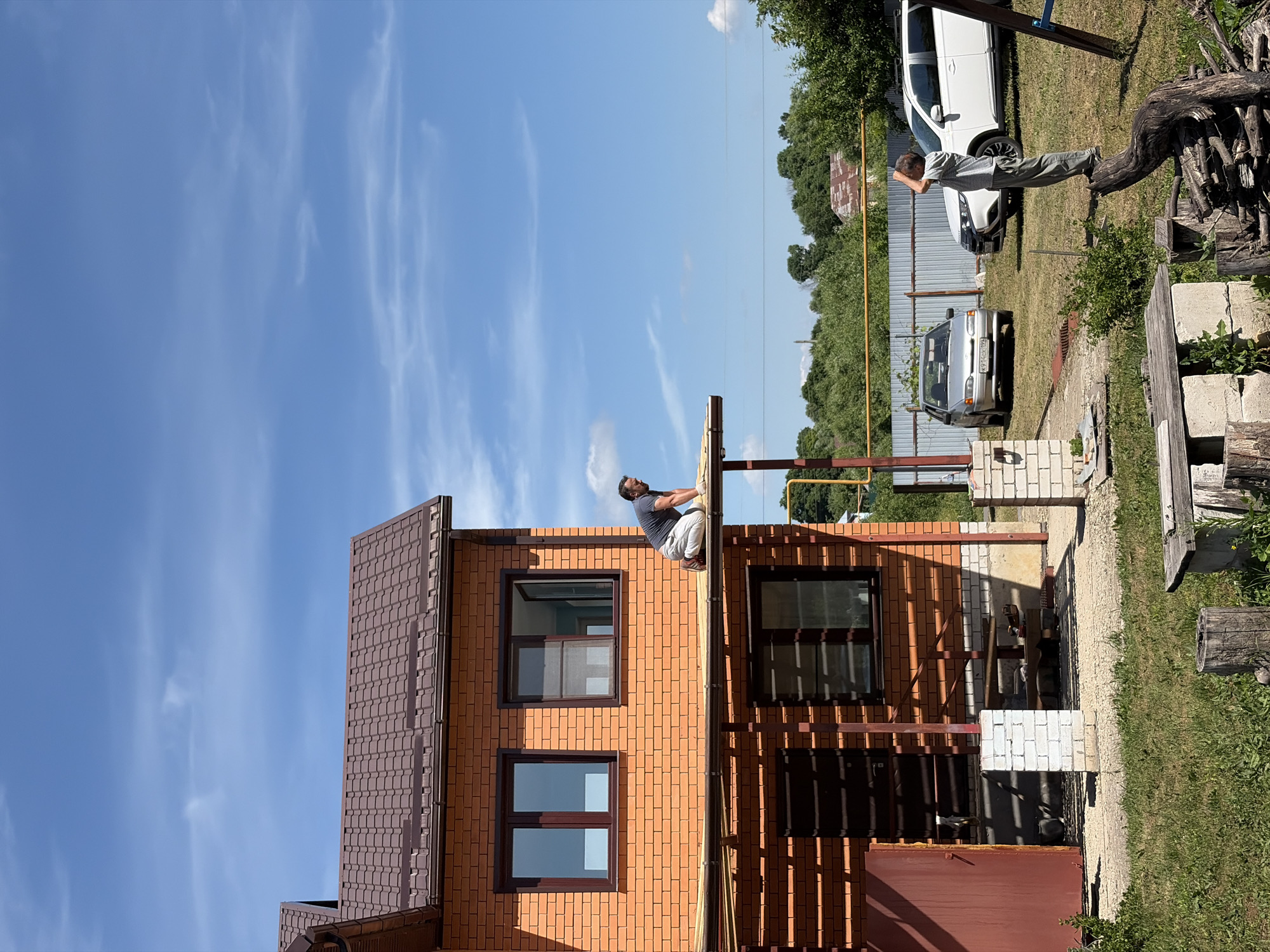 In Pavlovka, Russia, a man kneels on the edge of a partially constructed roof overhang of a two-story brick house. He is wearing a dark t-shirt, light-colored pants, and gloves. The house has a brown tiled roof and dark brown window frames. Below the overhang, there is a covered porch area with a red door and a window. In the yard, a young boy stands on a large piece of driftwood, looking up at the sky with his hand shielding his eyes. Two cars are parked in the grassy yard: a silver sedan and a white SUV. A metal fence runs along the background, behind which are trees and a partially visible rural structure. The sky is blue with scattered white clouds. The overall scene depicts outdoor construction and domestic life on a sunny day.