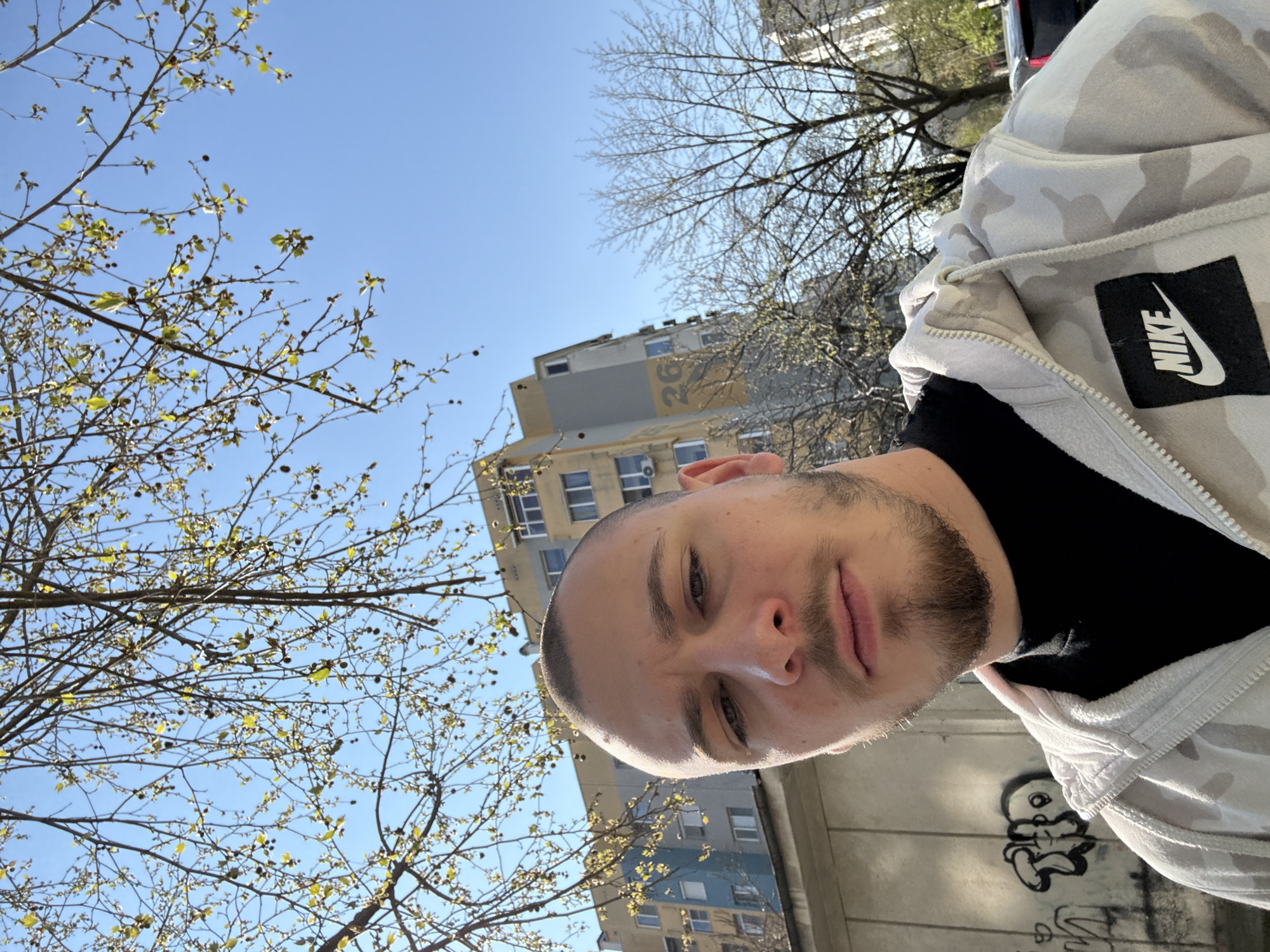 A medium shot of a Caucasian male in his late twenties taking a selfie outside. The sky is bright blue and there is a building visible in the background. The man is wearing a black shirt under a white and grey camouflage Nike hoodie. A tree with budding leaves is visible to the left. The graffiti on a wall behind the man is illegible, and there is a building located nearby. The location is Stara Zagora, Bulgaria.